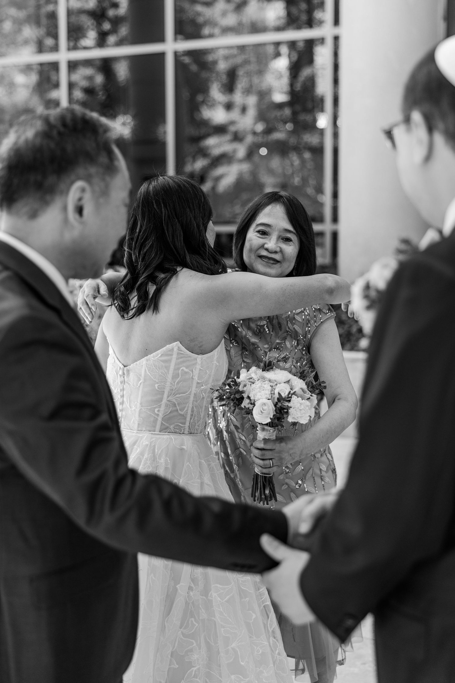 Black and white photo of a bride embracing a smiling woman holding a bouquet at a wedding ceremony.