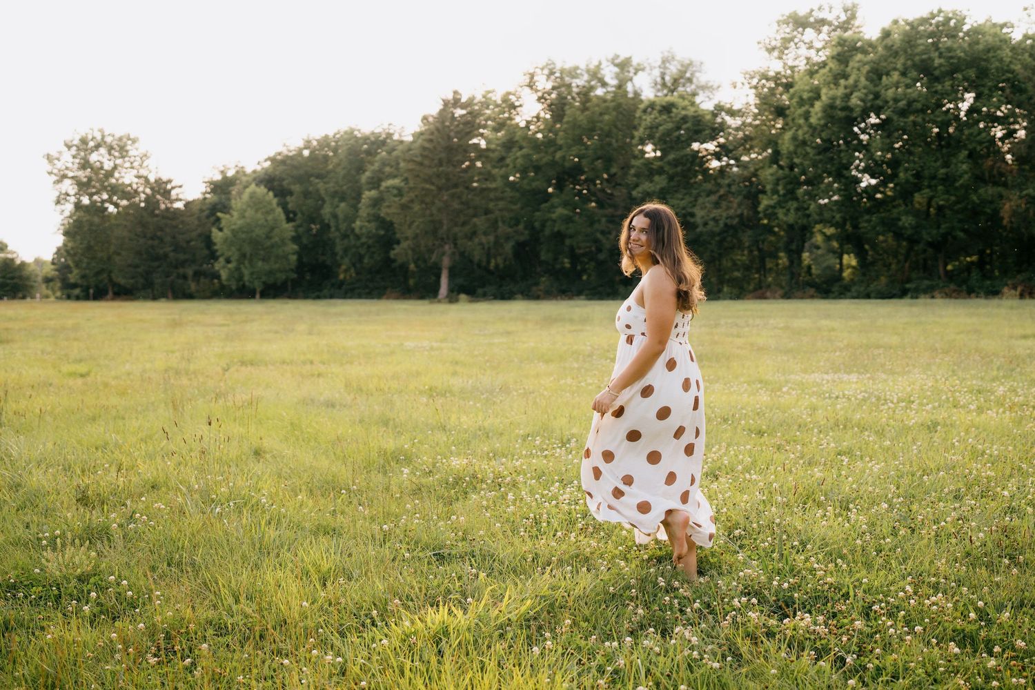 Woman in white polka dot sundress walking through a sunny green meadow with trees in the background.