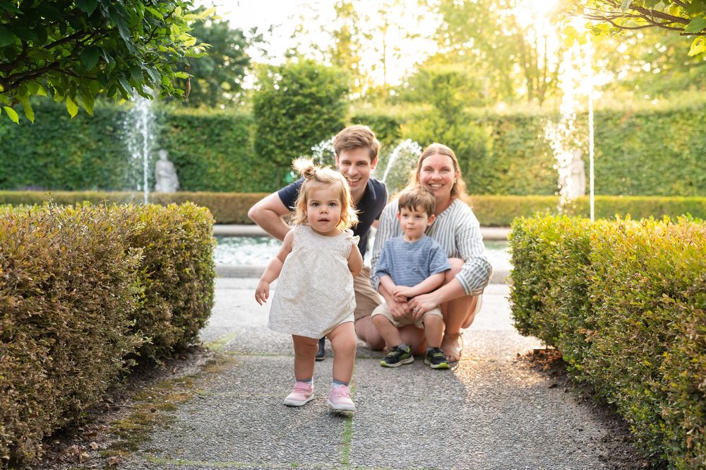 Family of four poses in a lush garden path with fountains, parents crouching behind two young toddlers in golden light.