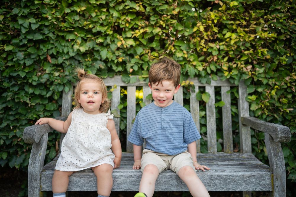 Two young children, a toddler girl and a boy, sit smiling on a wooden garden bench surrounded by lush green ivy.