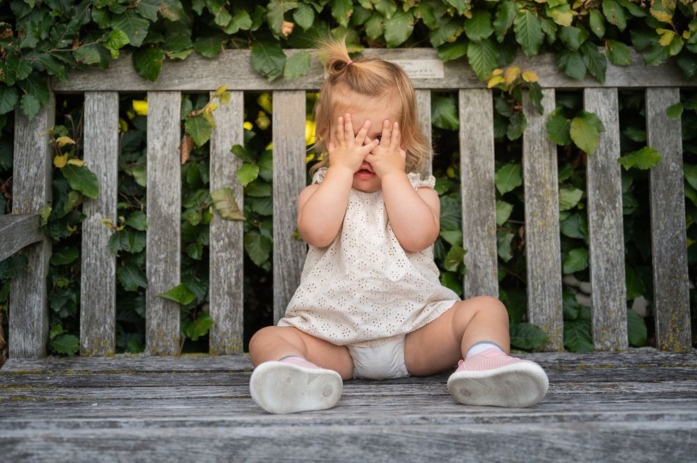Toddler girl in white romper covering her face with hands while sitting on a wooden bench outdoors.