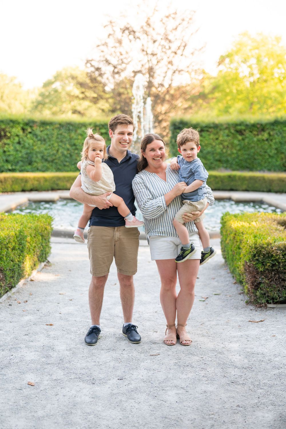 Family of four posing in a garden with fountain, parents holding two toddlers, surrounded by trimmed hedges.