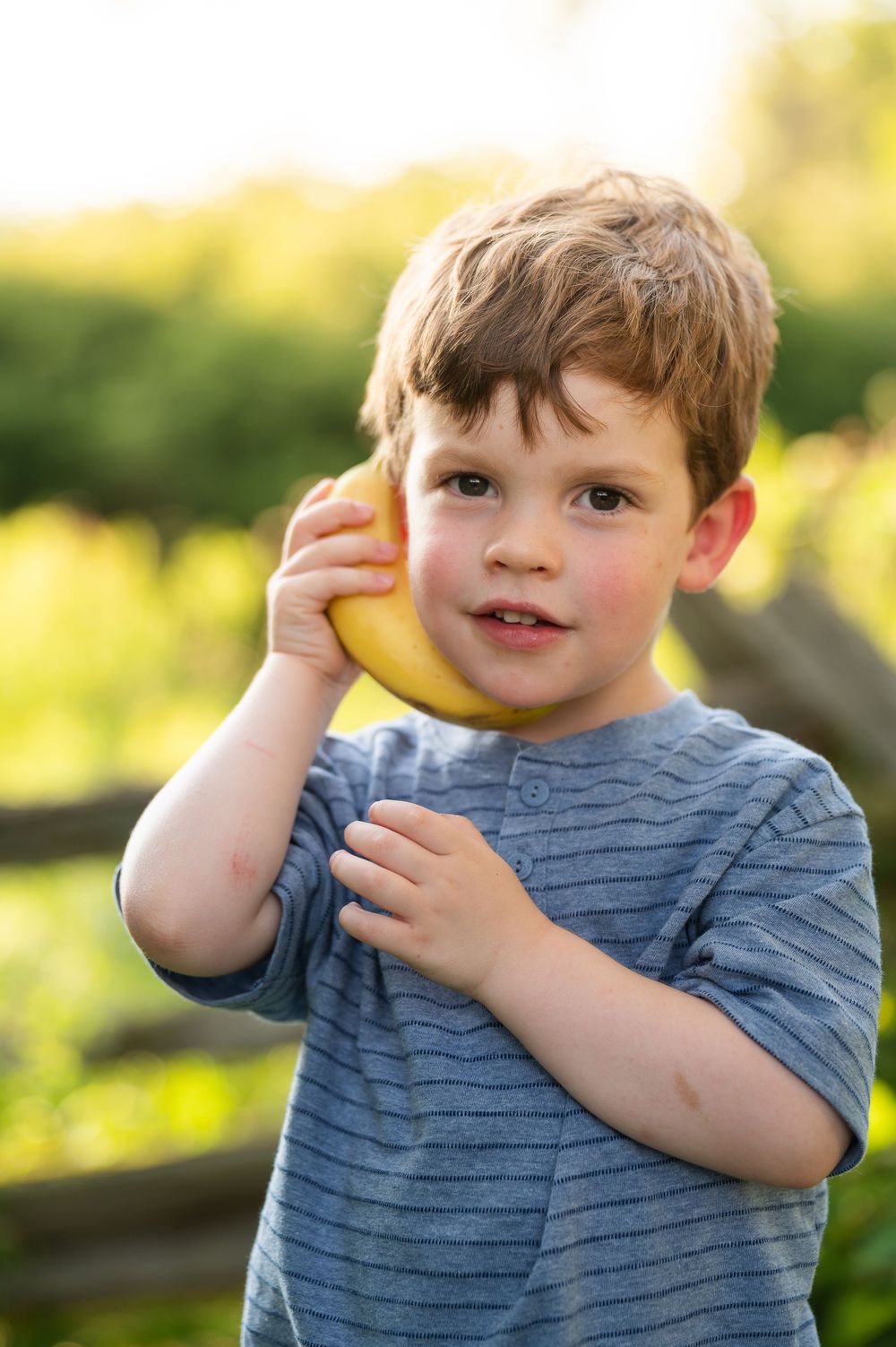 Young boy holds a banana like a phone to his ear outdoors, smiling playfully in natural sunlight.