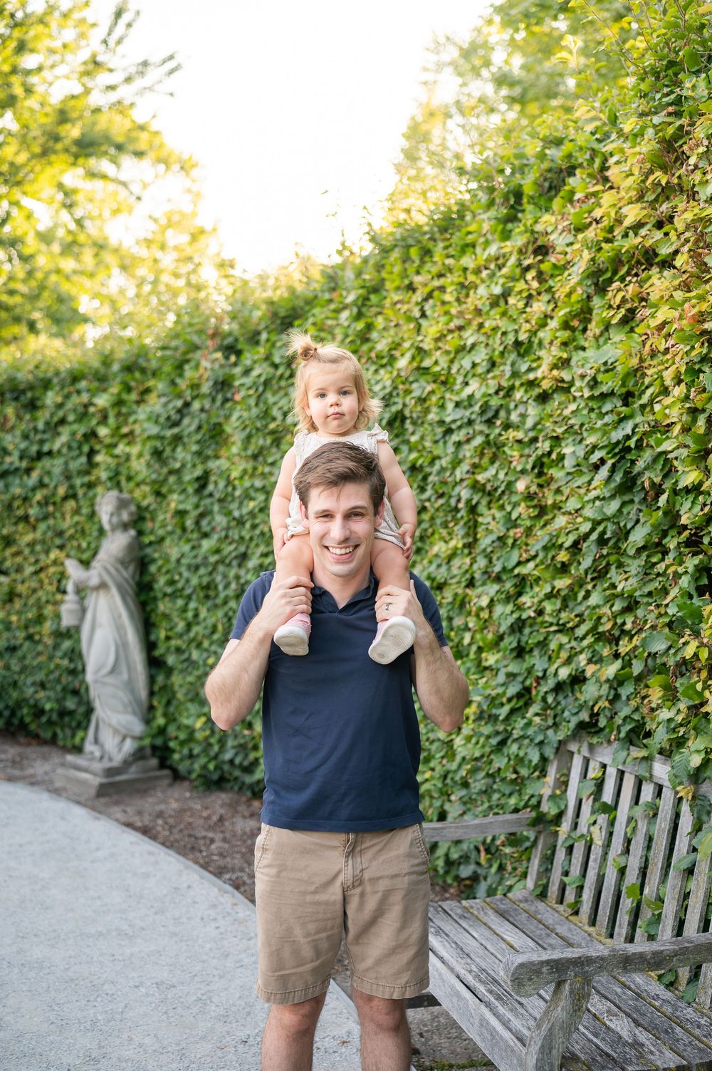 A smiling man carries a toddler on his shoulders in a lush garden with ivy walls and a stone statue.