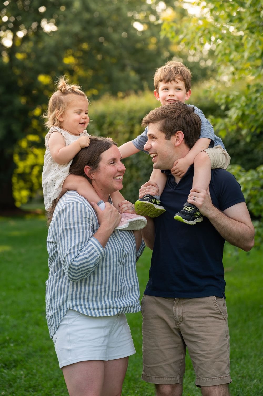 Happy family of four outdoors, parents carrying young children on shoulders, smiling in a lush green park setting.