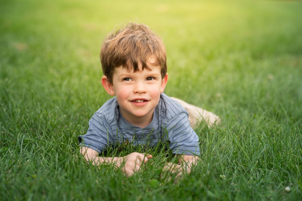 Happy young boy lying on lush green grass, smiling at camera on a sunny day outdoors.