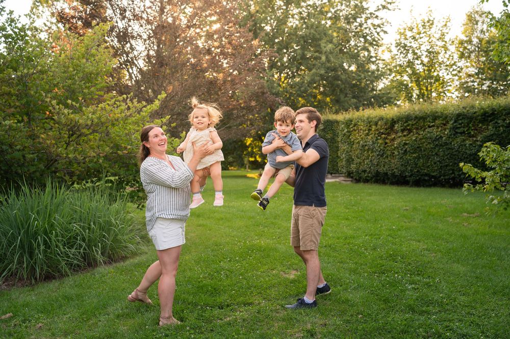 Family of four playing outdoors in lush green garden, parents lifting two young children in the air during golden hour.