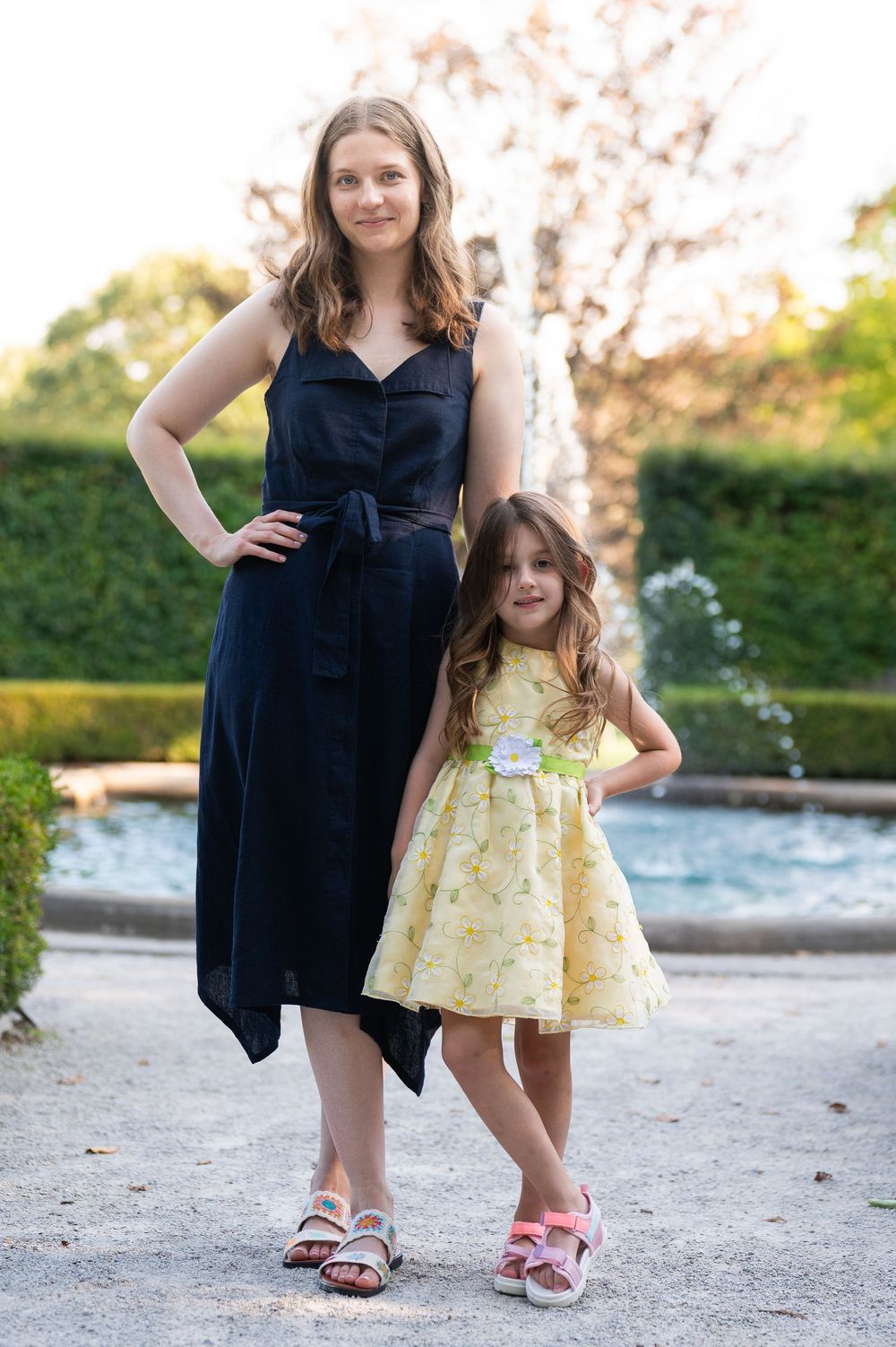 Woman in navy dress and young girl in yellow floral dress posing together outdoors near a fountain.