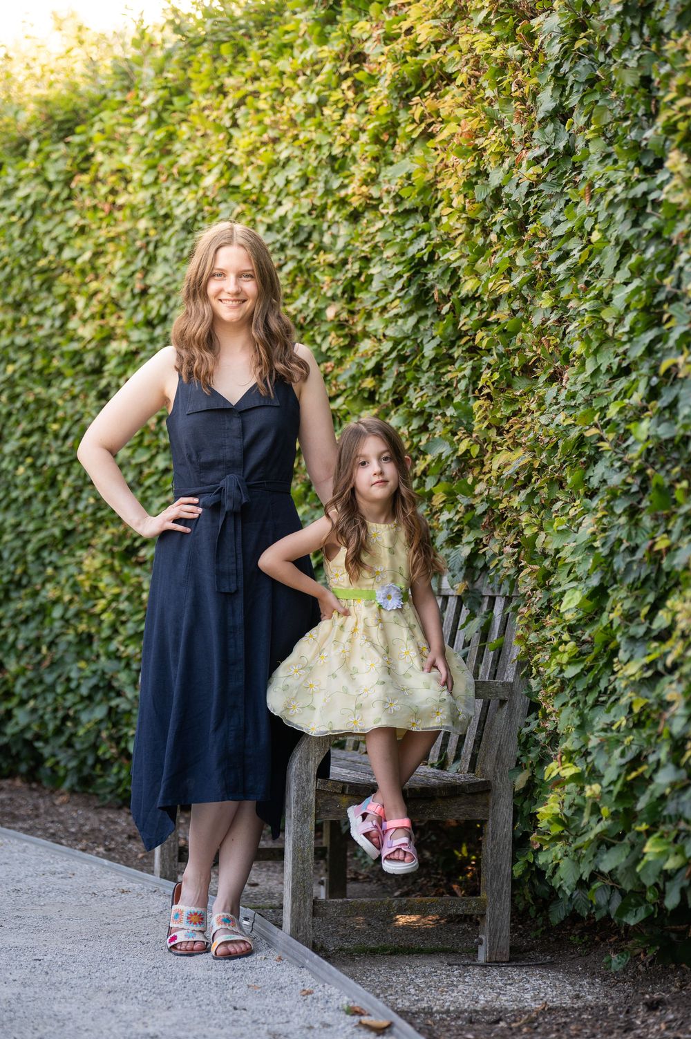 Woman in navy dress and girl in yellow dress pose by a wooden bench against a lush green ivy wall outdoors.