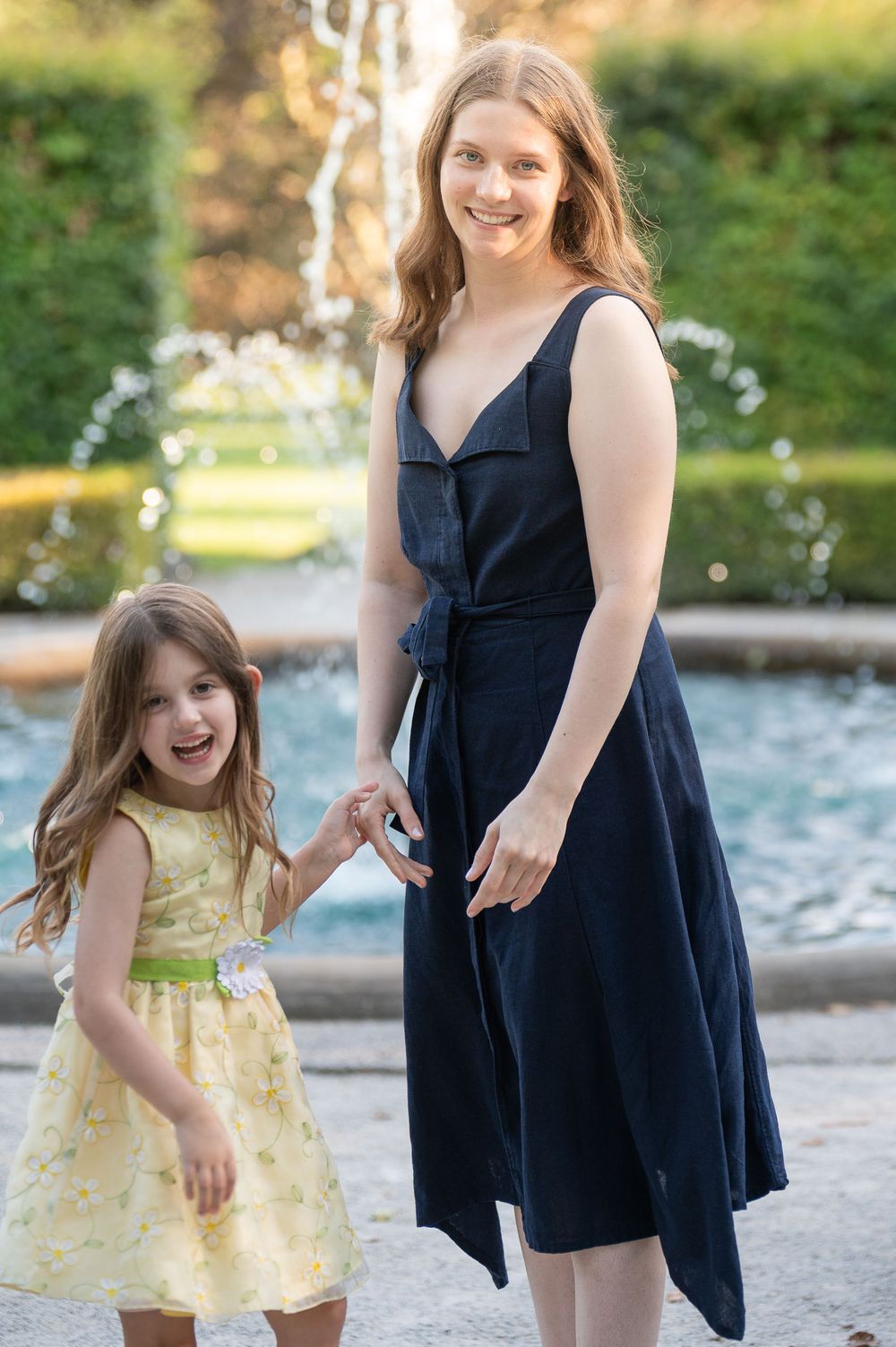 Woman in navy dress holds hands with young girl in yellow floral dress near garden fountain.