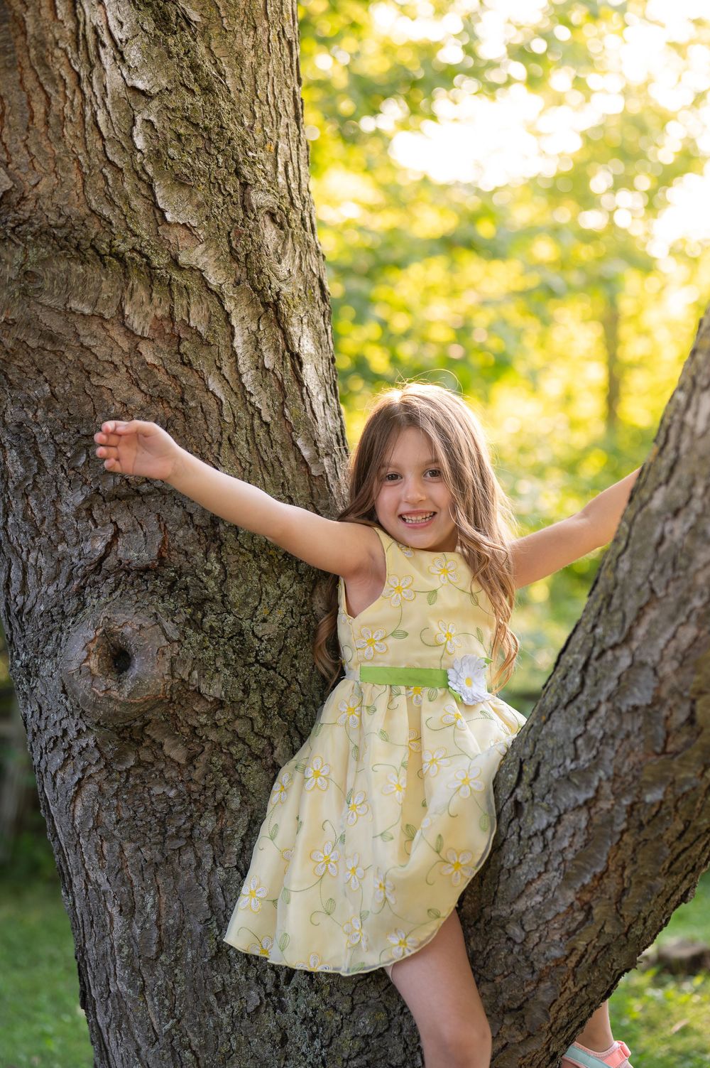 Young girl in yellow floral dress smiling while leaning against a large tree trunk in a sunny park.