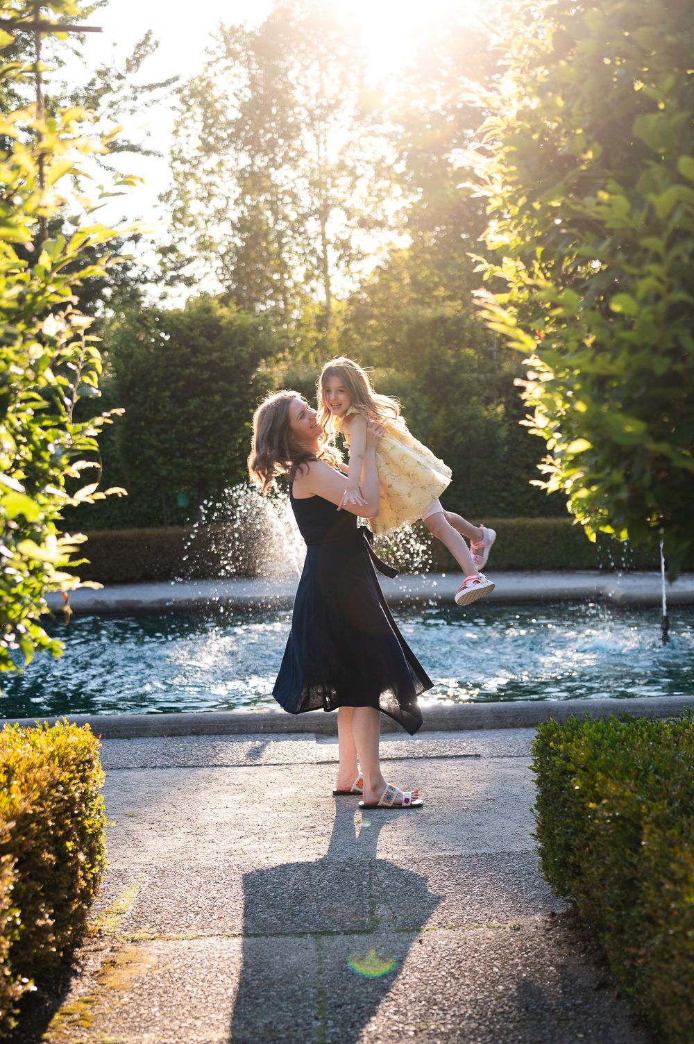 Mother in black dress lifts smiling daughter in yellow dress by sunlit garden fountain.