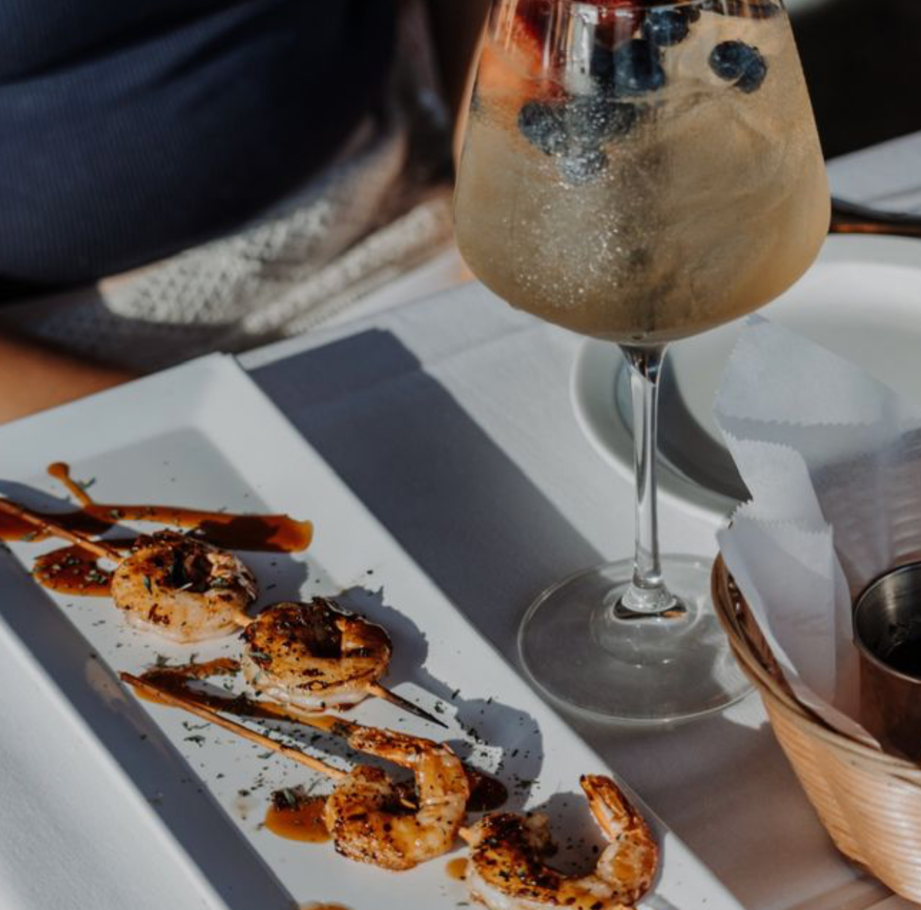 Grilled shrimp appetizer with drizzle sauce on white plate beside a sparkling cocktail with blueberries.