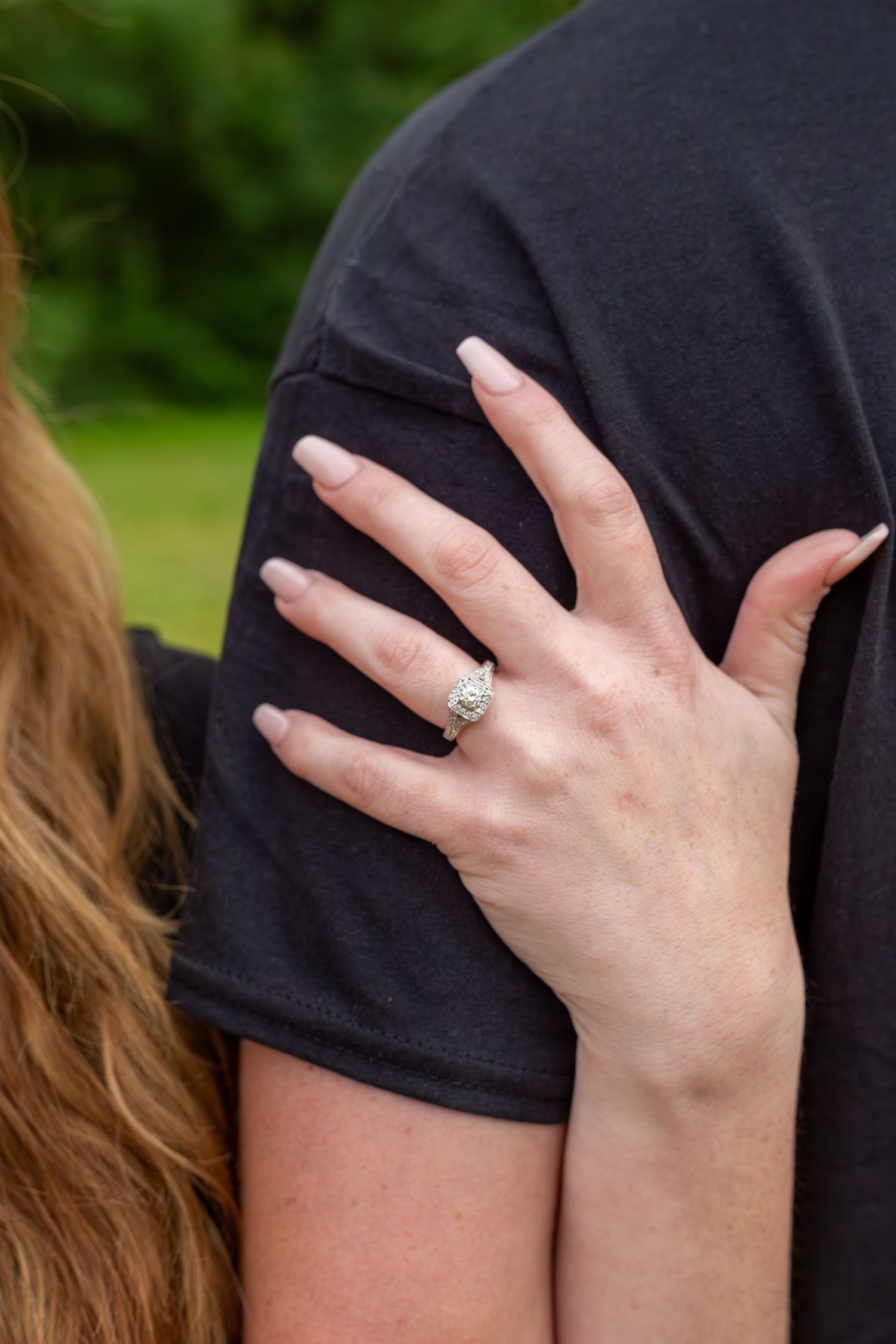 Woman displaying diamond engagement ring on finger while resting hand on mans shoulder outdoors.