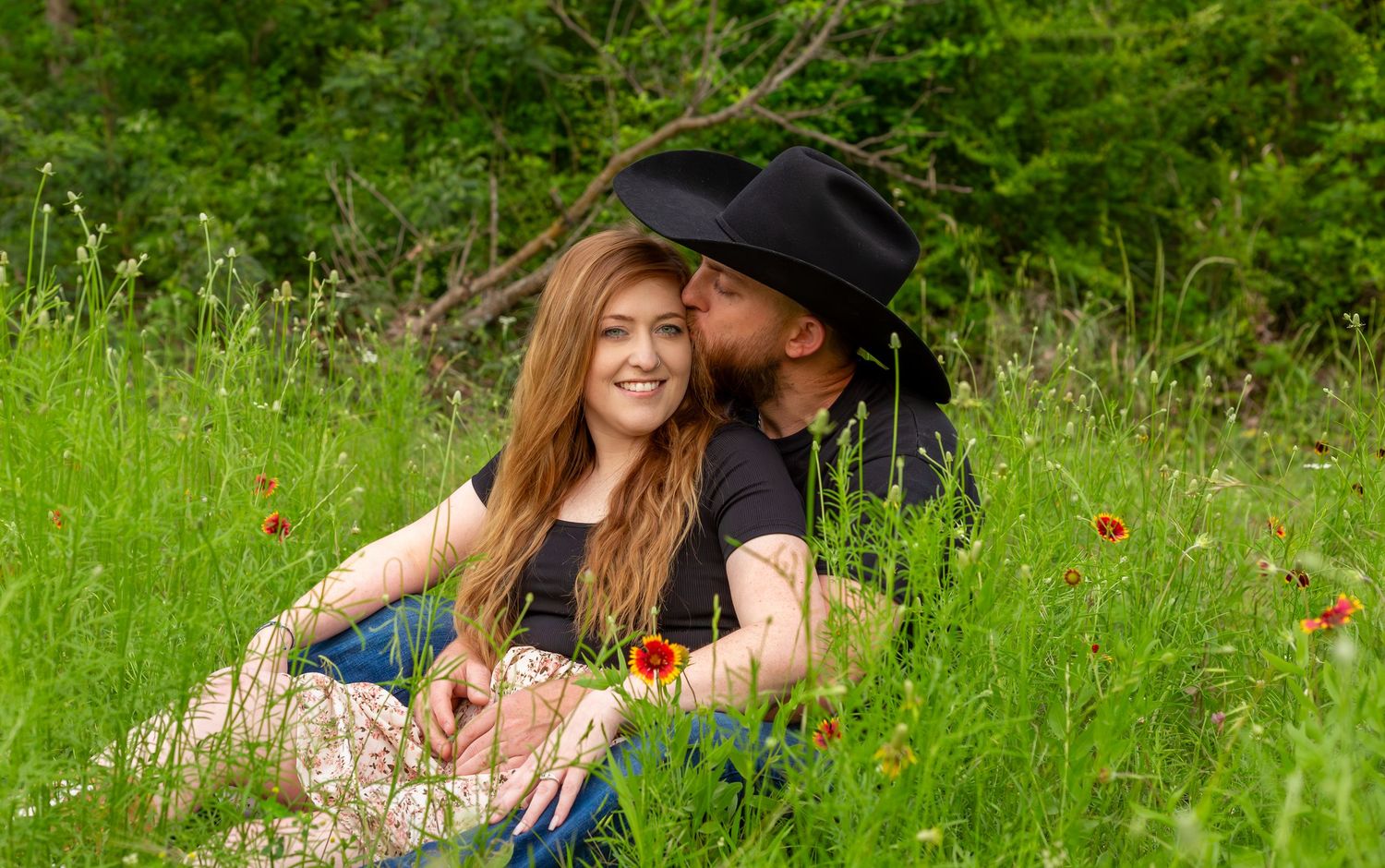 A couple sits in a lush green meadow with wildflowers, sharing a romantic kiss outdoors.