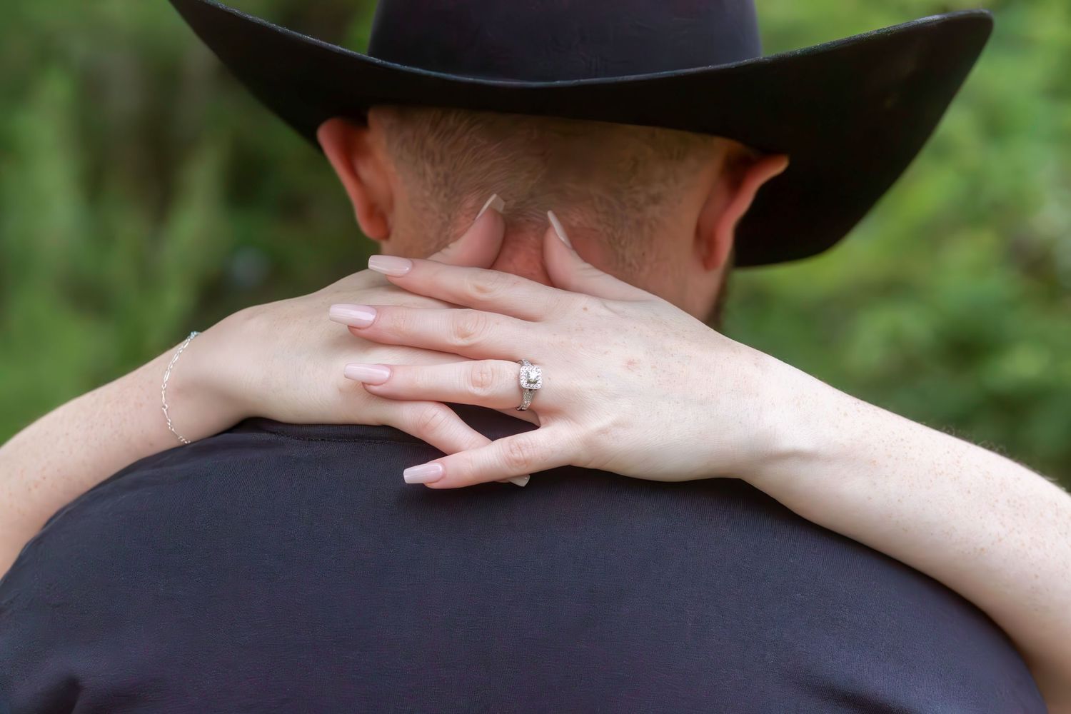 Woman with engagement ring resting her hand on a man's shoulder wearing a black cowboy hat outdoors.