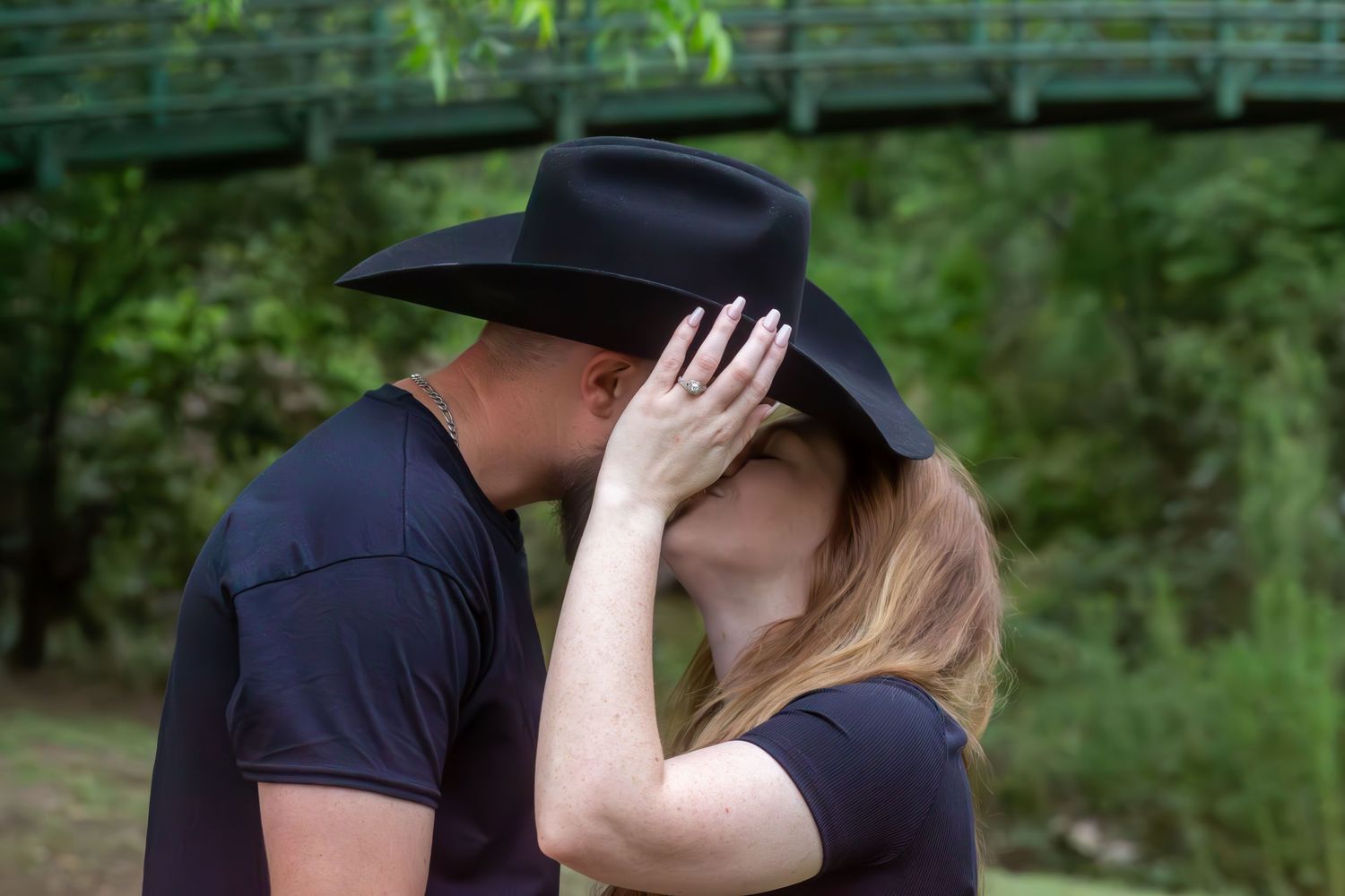 Couple sharing a romantic kiss outdoors, woman wearing a black cowboy hat, with a green bridge in the background.