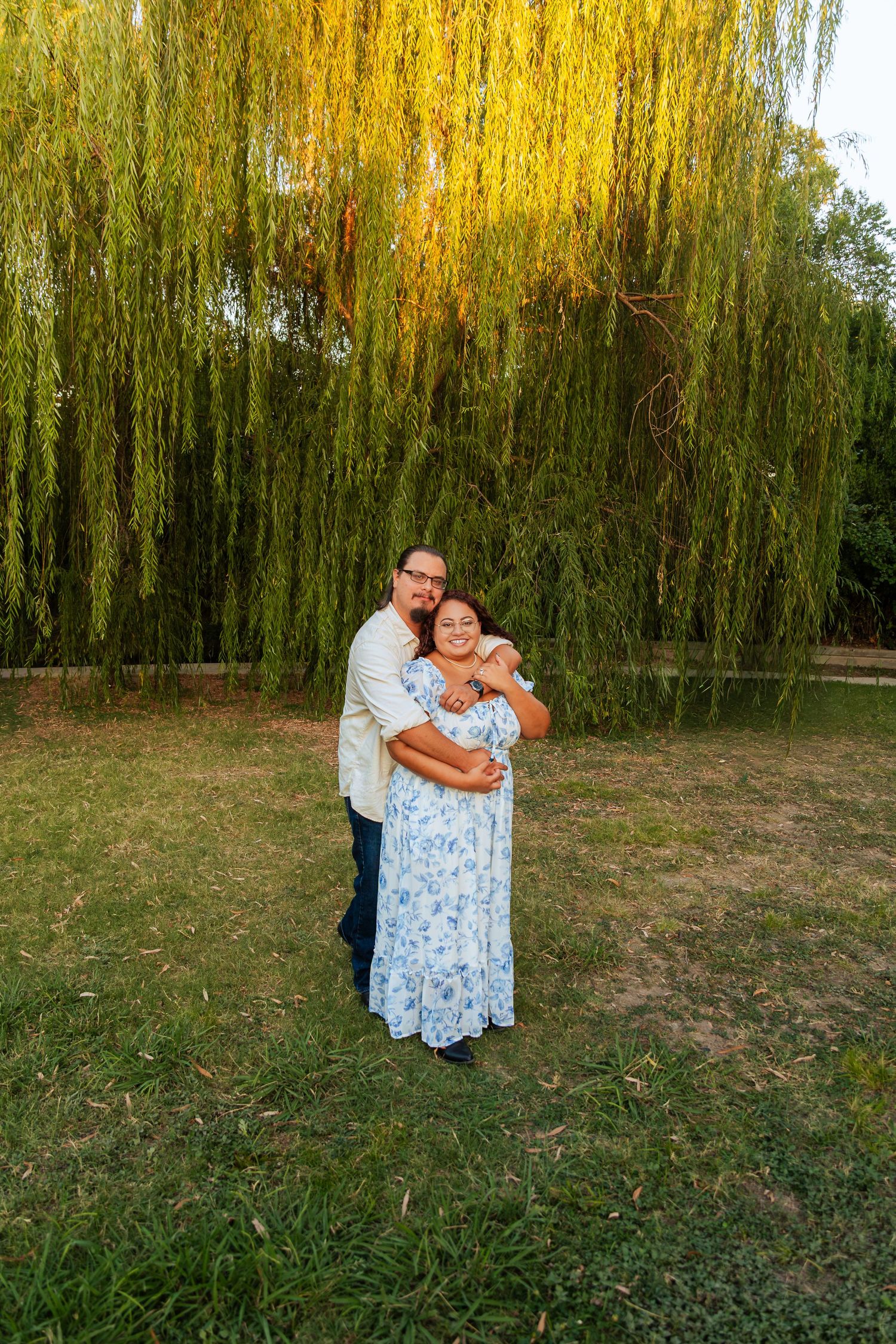 Couple embracing under a weeping willow tree on green grass during golden hour outdoors.