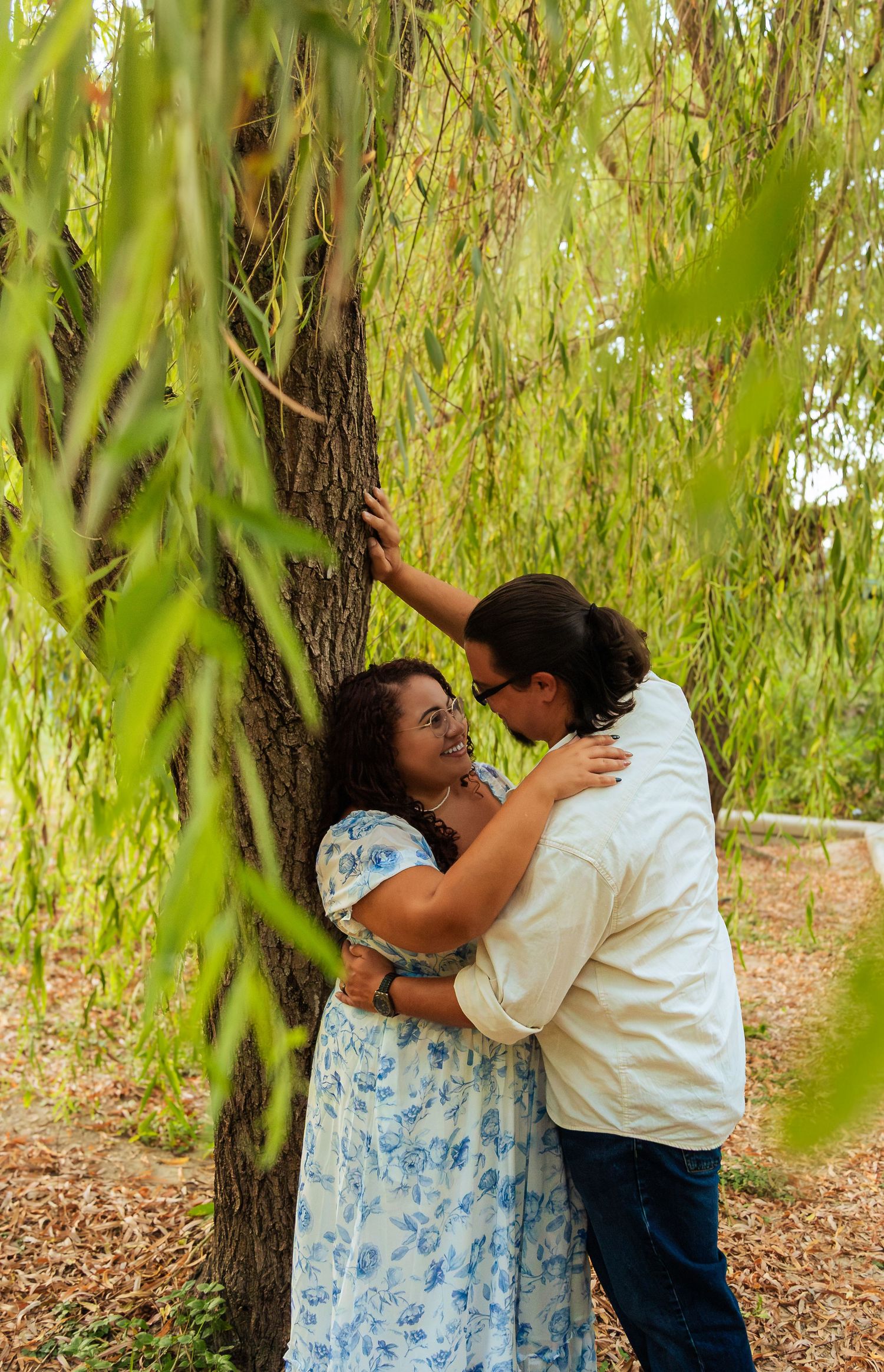 Couple embracing romantically under a weeping willow tree, surrounded by lush green foliage outdoors.