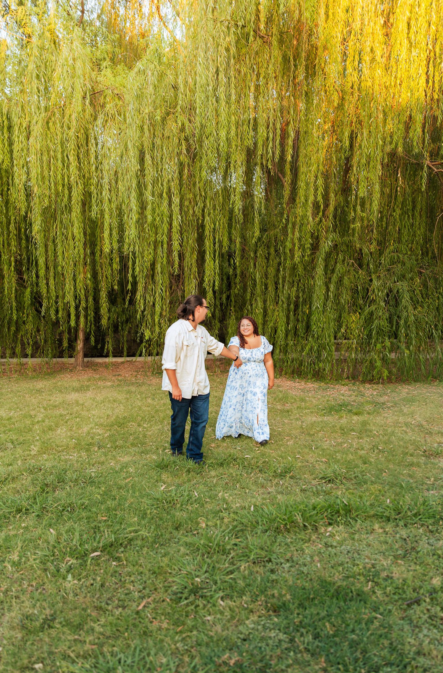 Couple holding hands and dancing on green grass in front of a large weeping willow tree at golden hour.