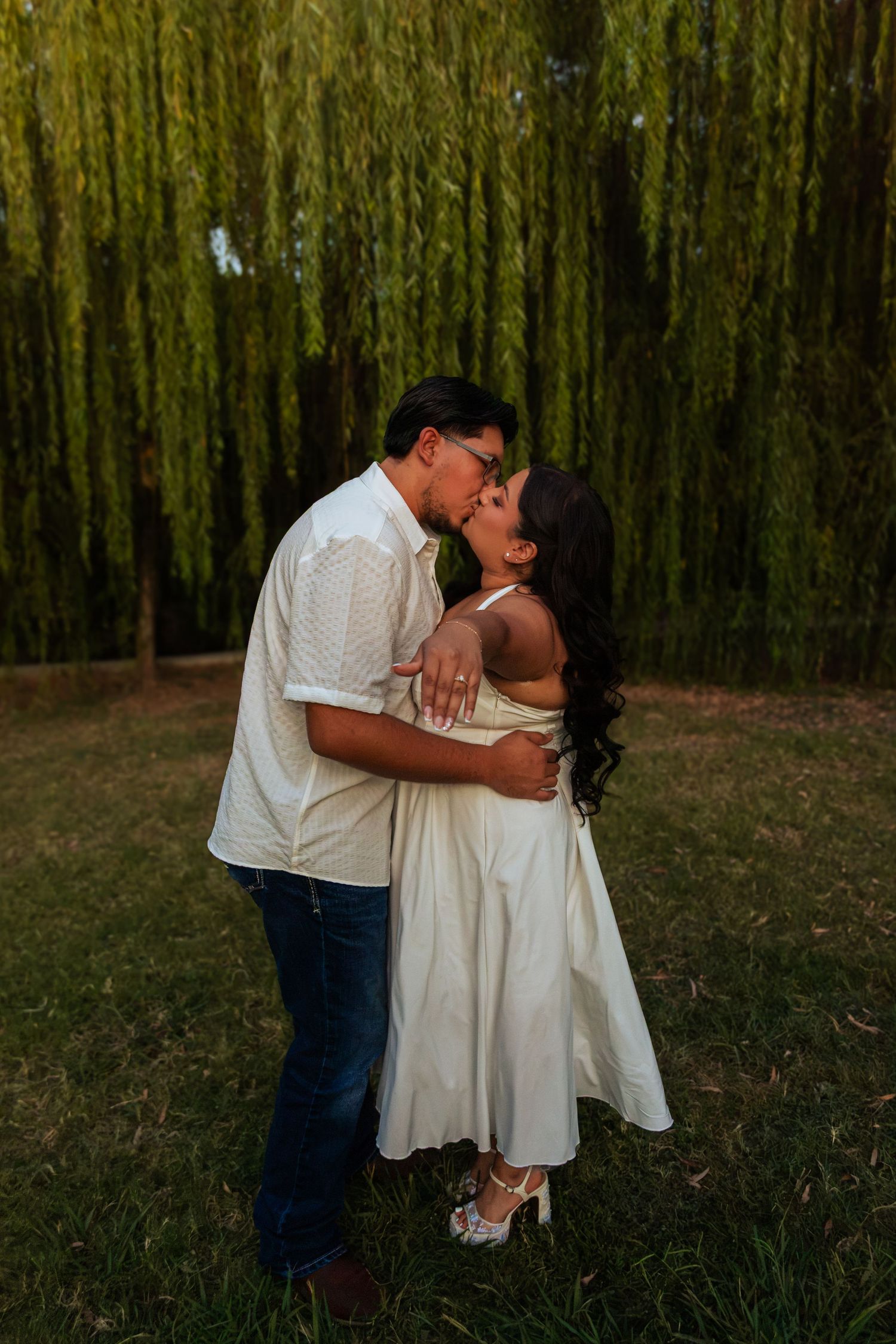 Couple sharing a romantic kiss outdoors in front of a weeping willow tree, wearing white outfits in golden hour light.