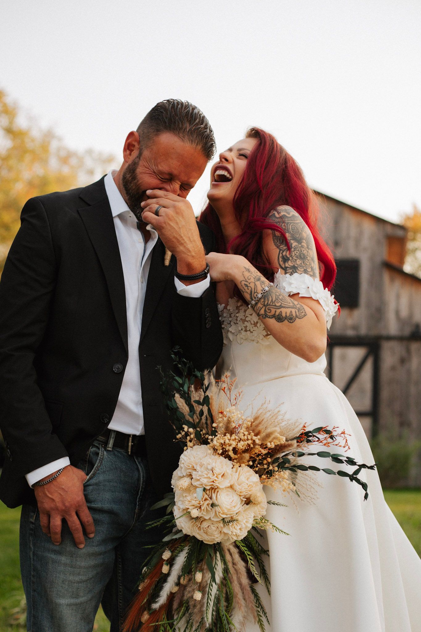 Laughing bride with red hair and tattoos celebrates with groom holding a boho wedding bouquet.