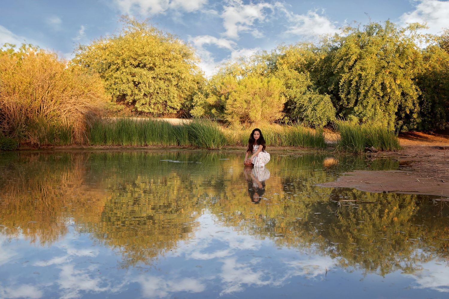 Woman in a white dress sits at the edge of a calm lake, her reflection mirrored in the water beside golden autumn trees.