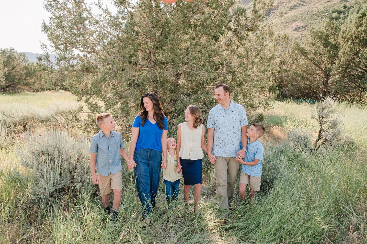 Family of six walking together on a nature trail surrounded by green shrubs and trees on a sunny day.