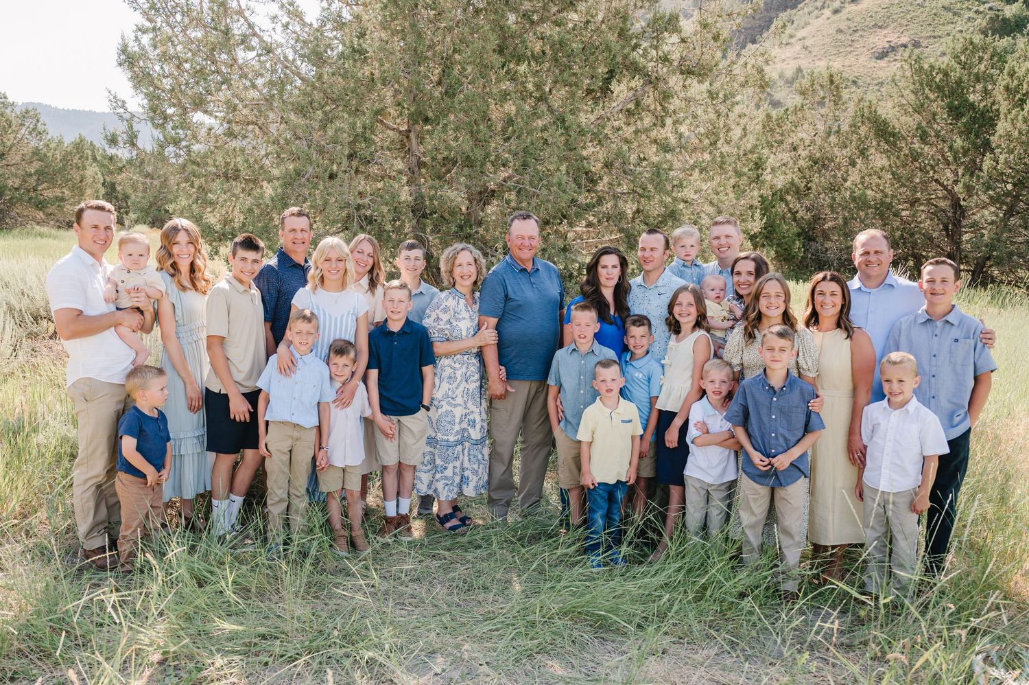 Large multigenerational family portrait outdoors in a meadow with green trees, wearing coordinated neutral and blue outfits.