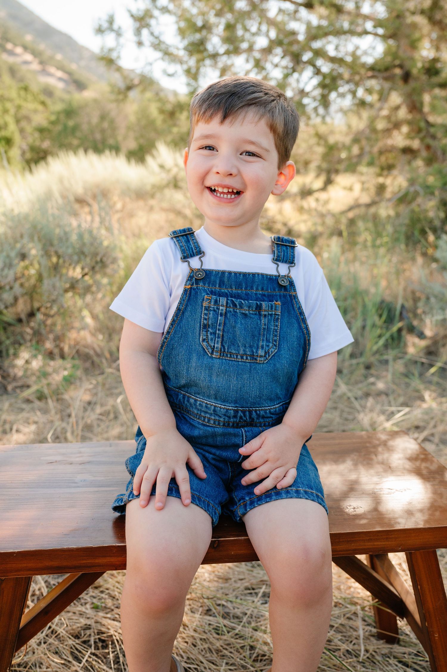 Smiling young boy in denim overalls and white t-shirt sitting on wooden bench outdoors in natural setting.