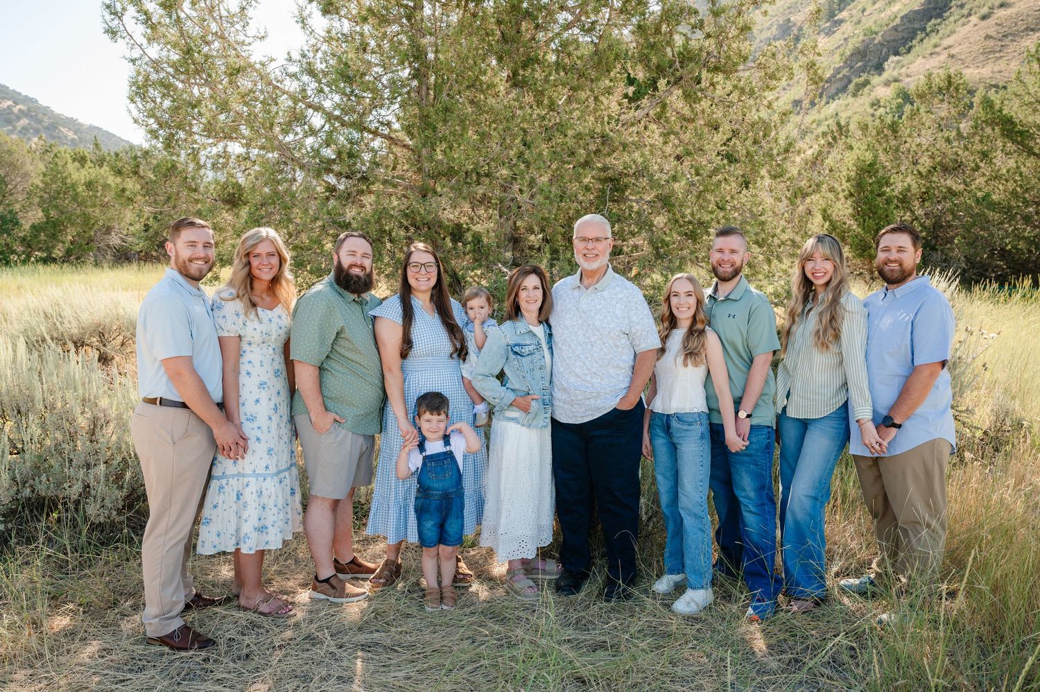 Large family group portrait outdoors in a meadow with mountains and trees in the background on a sunny day.