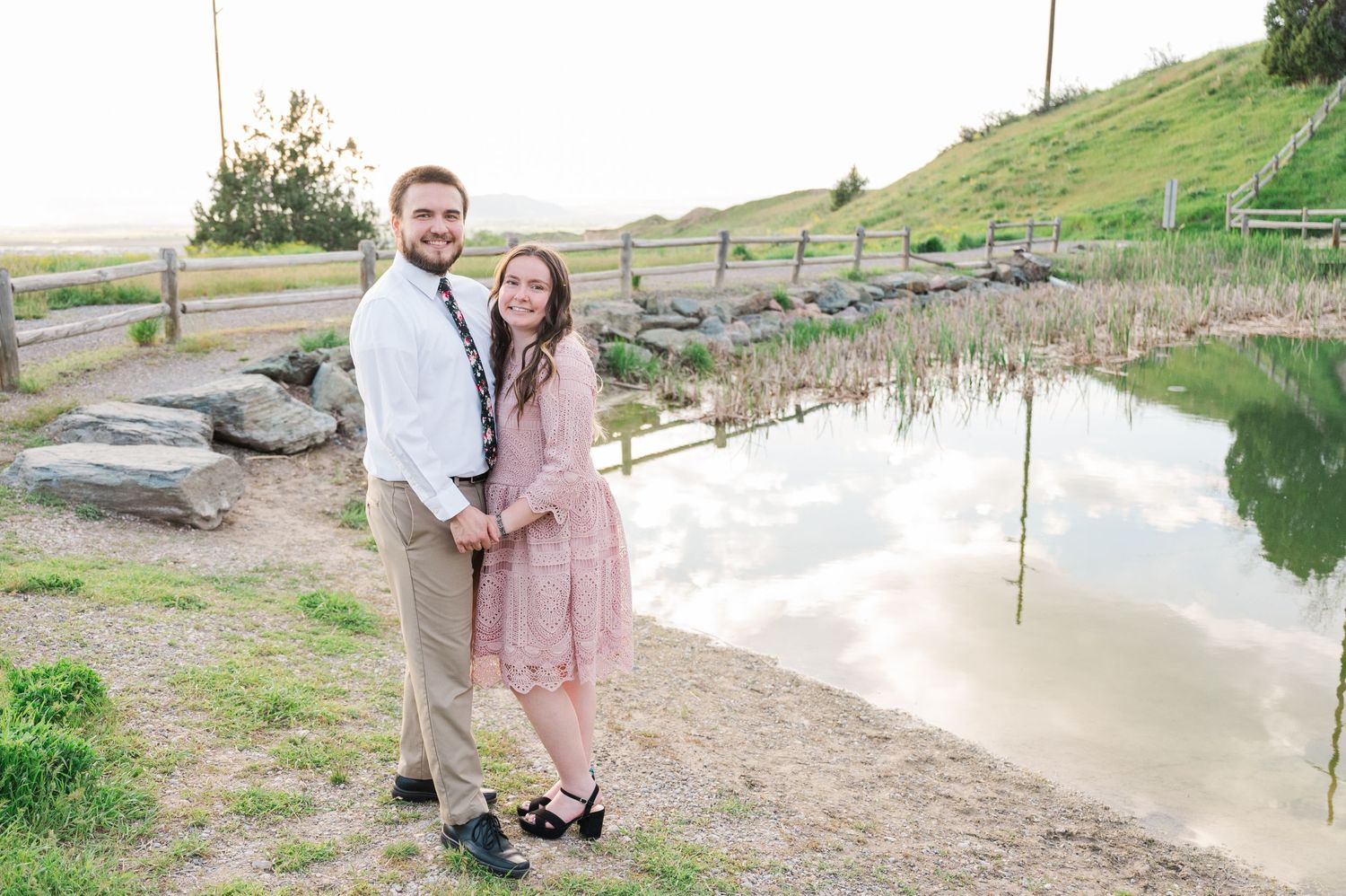 A couple poses together by a tranquil pond with hills in the background.