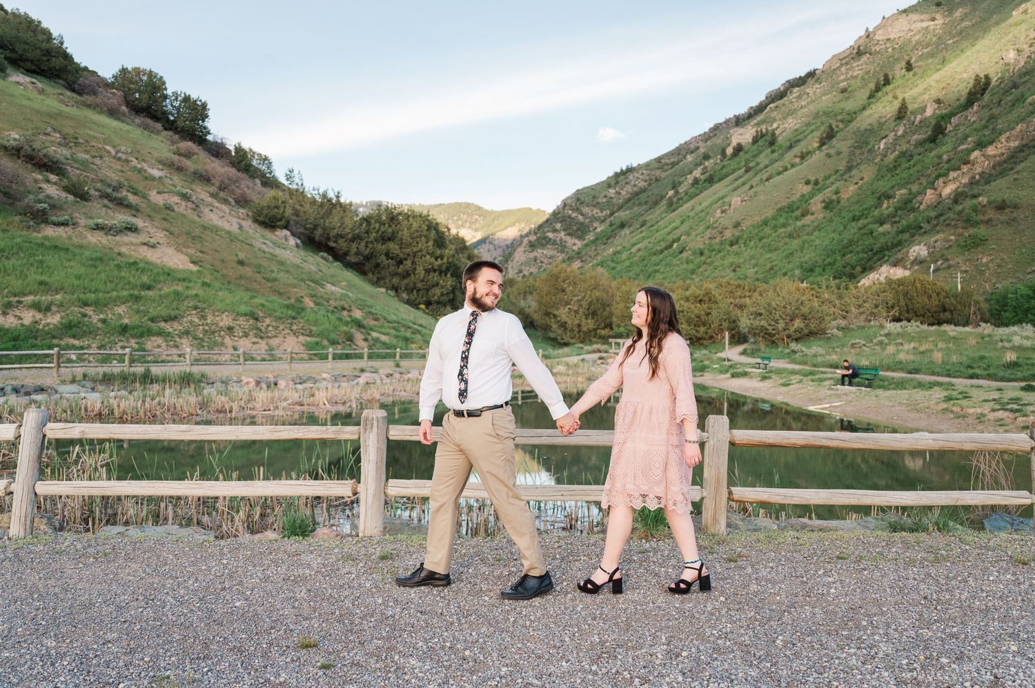 A couple holds hands while posing on a gravel path with rolling hills behind them.