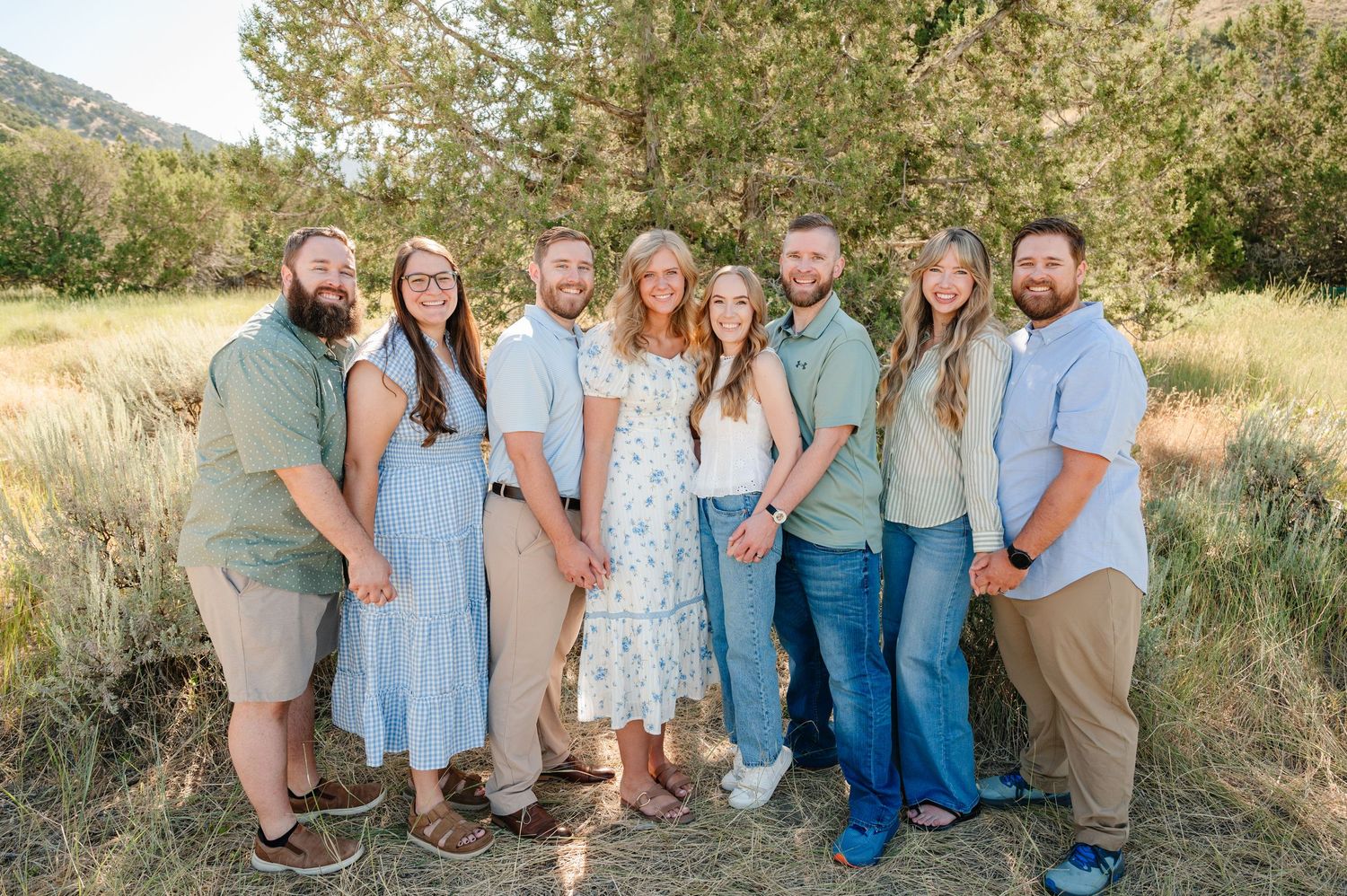 A joyful group of eight adults posing together outdoors in a natural field setting with trees in the background.