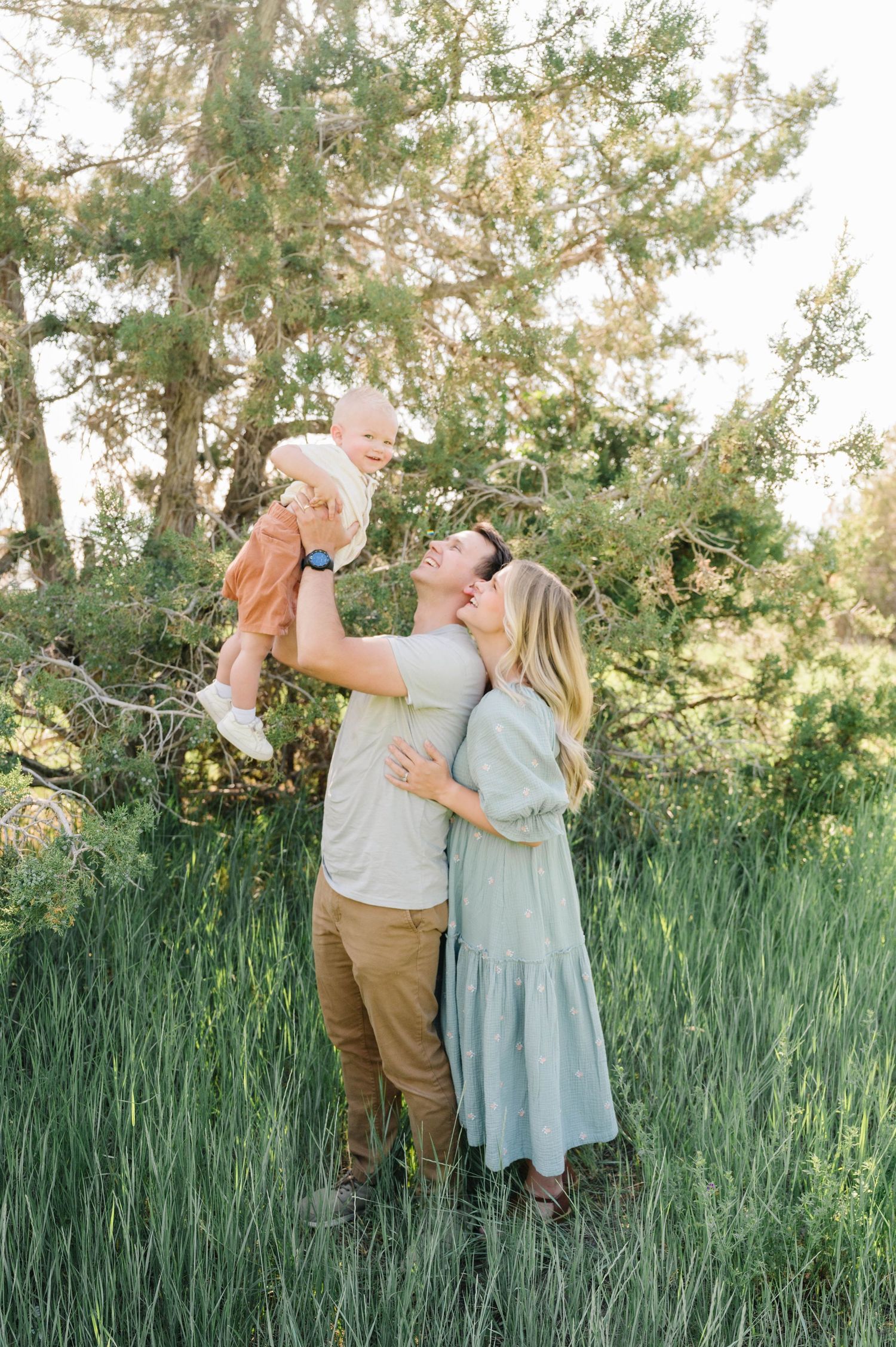 A family shares a joyful moment together in tall summer grass with evergreen trees behind them.
