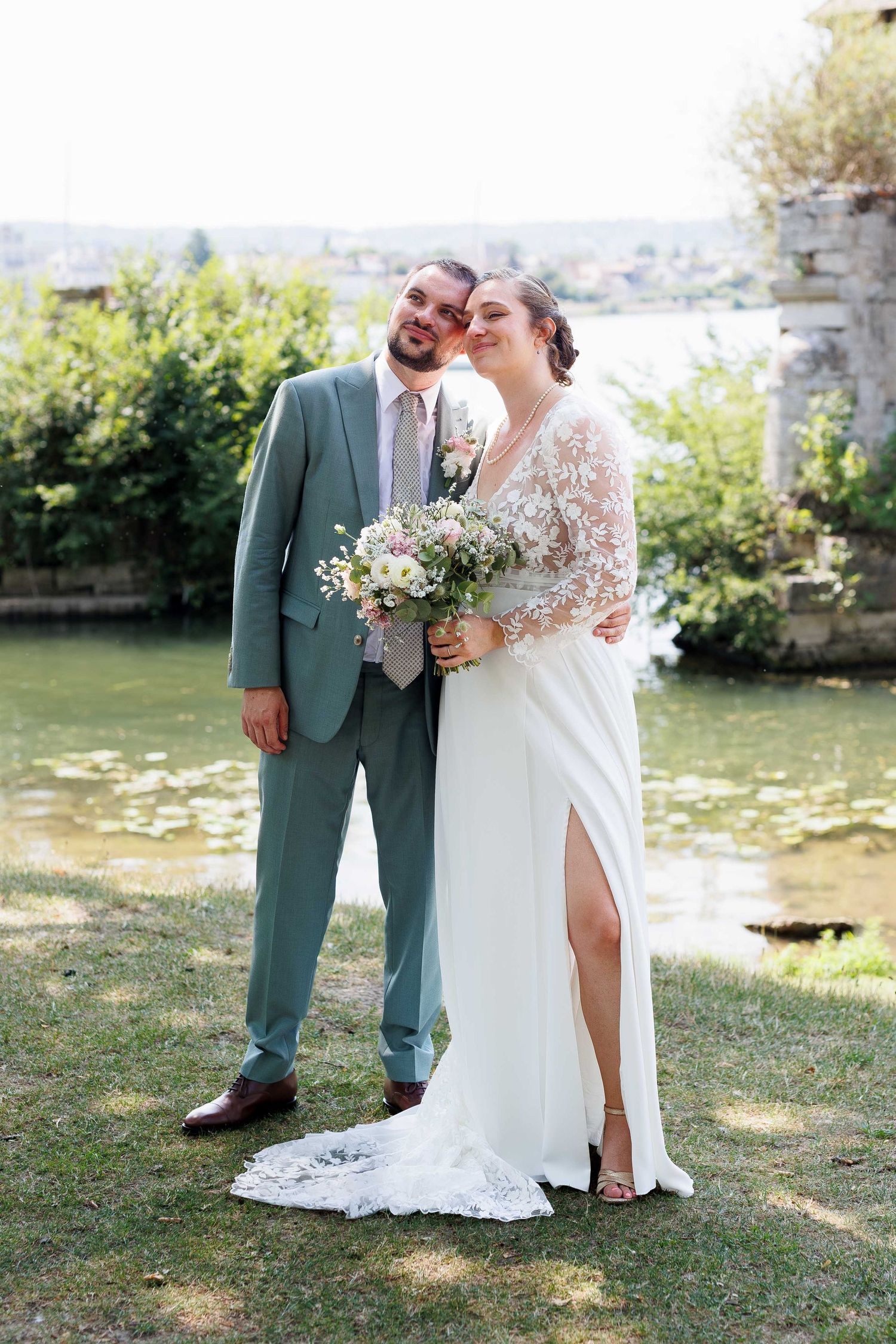 Bride and groom pose by a pond, she holds a bouquet in a white gown, he wears a sage green suit.