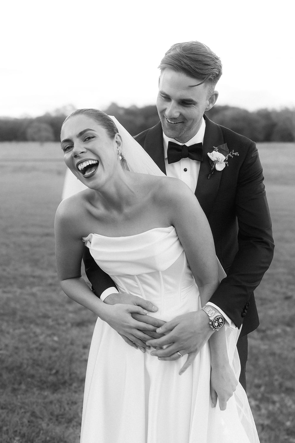 Black and white photo of a laughing bride in strapless gown embraced by smiling groom in tuxedo outdoors.