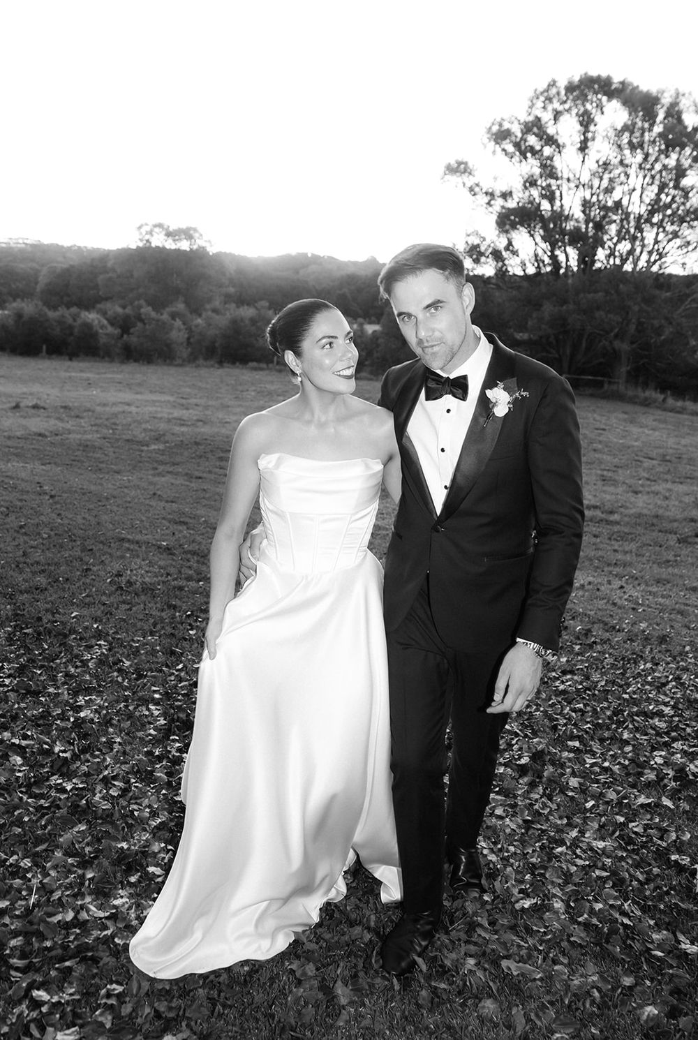 Black and white photo of a bride in strapless gown and groom in tuxedo posing outdoors at their wedding.