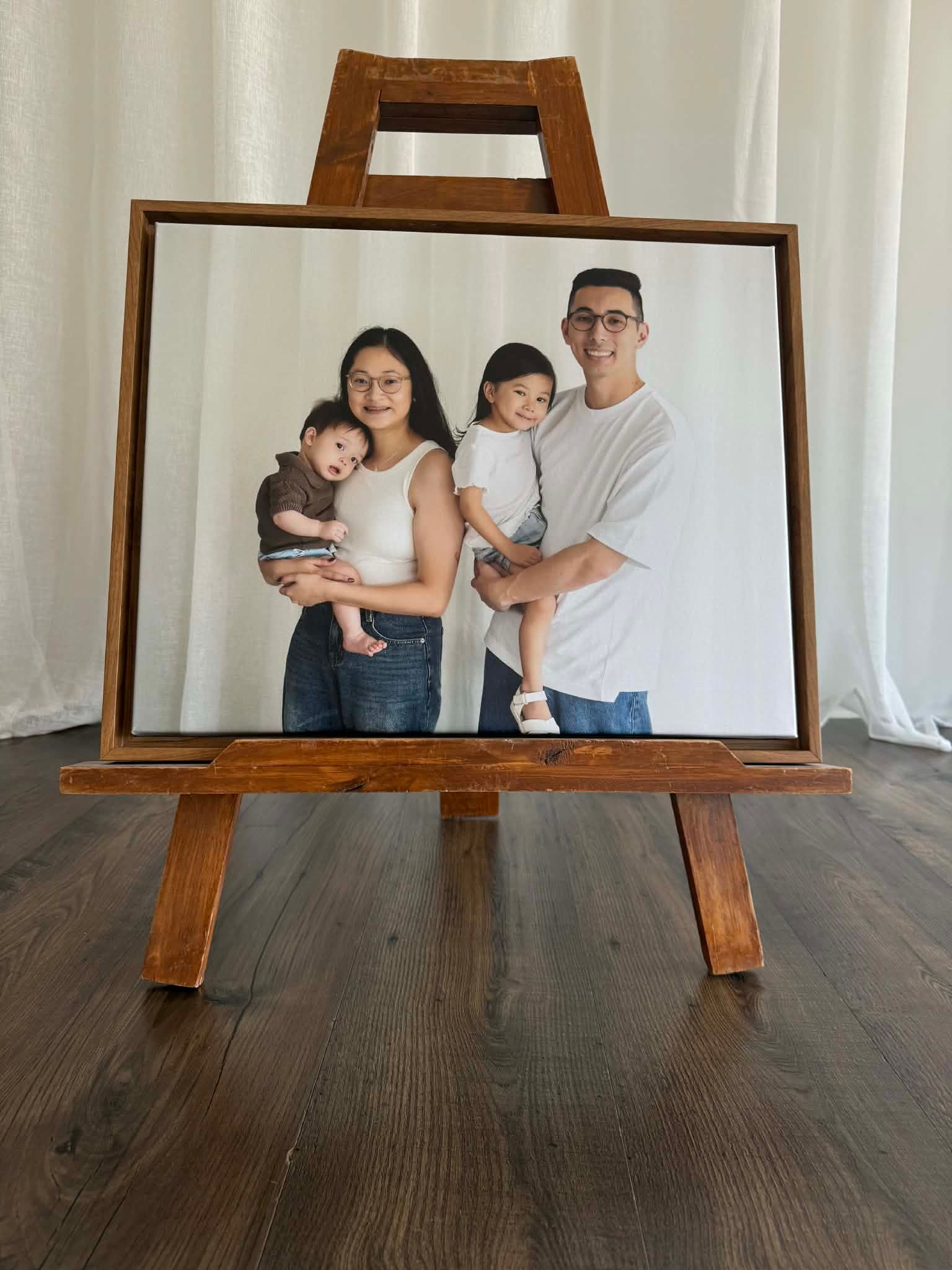 Framed family portrait on wooden easel showing parents holding two young children against white background.
