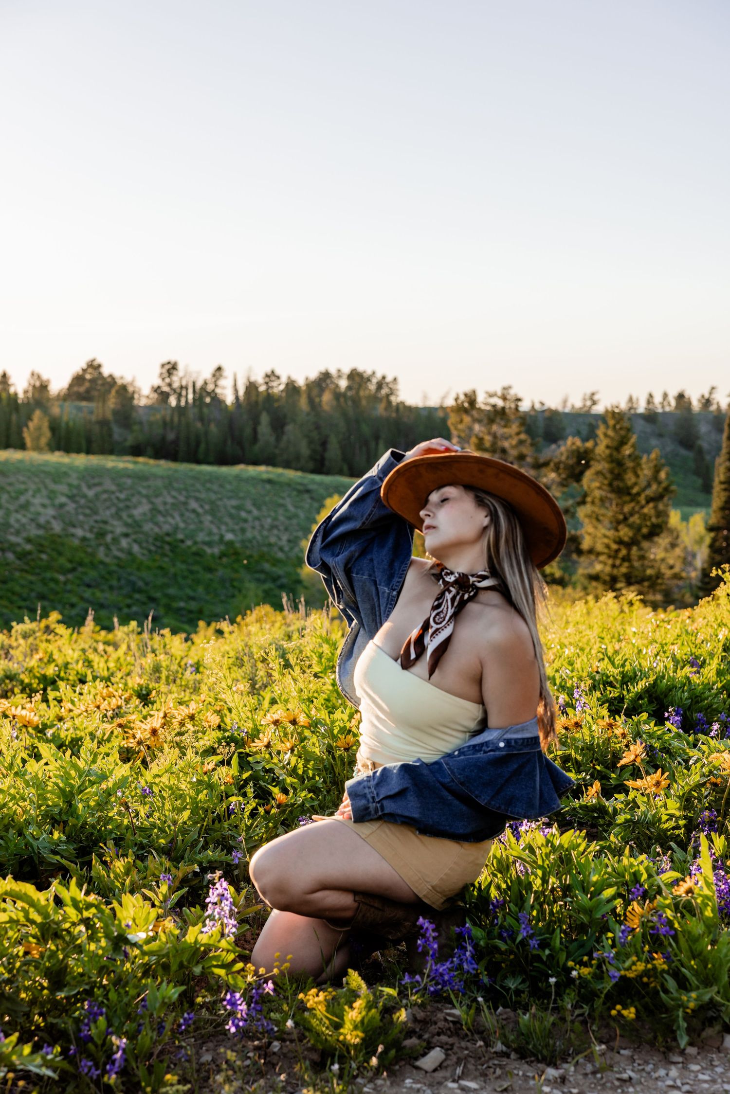 Woman in cowboy hat and denim jacket poses among wildflowers on a sunlit hillside with evergreen trees in background.