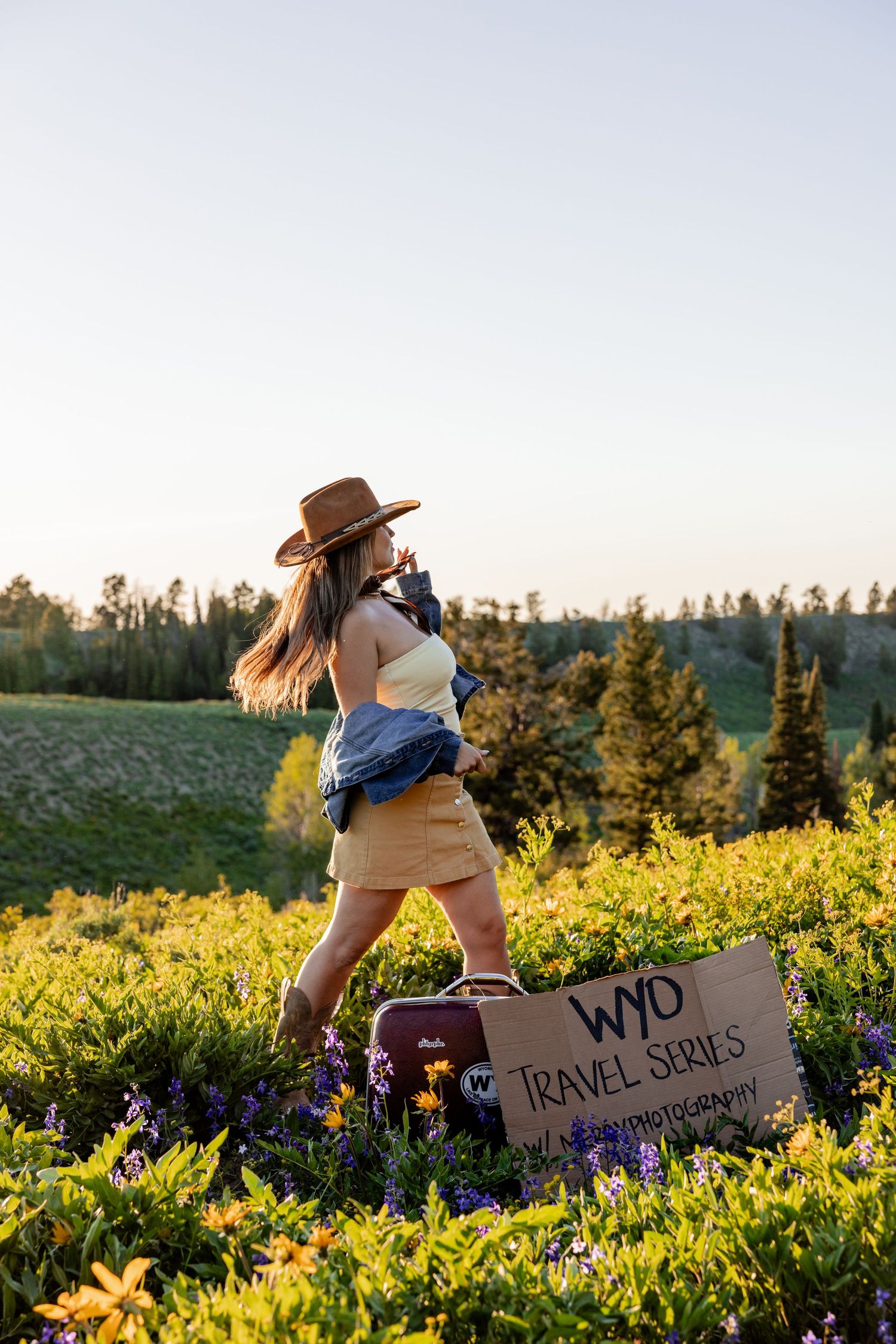 Woman in cowboy hat walking through wildflower meadow beside a WYO Travel Series cardboard sign at golden hour.