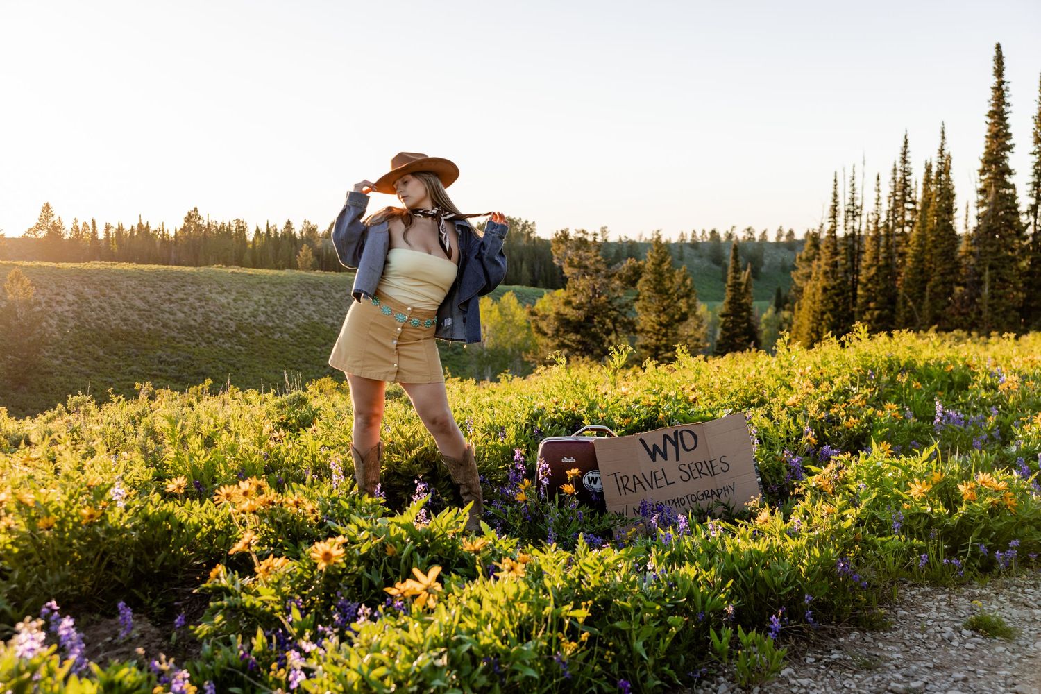 Hiker in hat and denim jacket stands among wildflowers at golden hour near a trail sign in a mountain meadow.