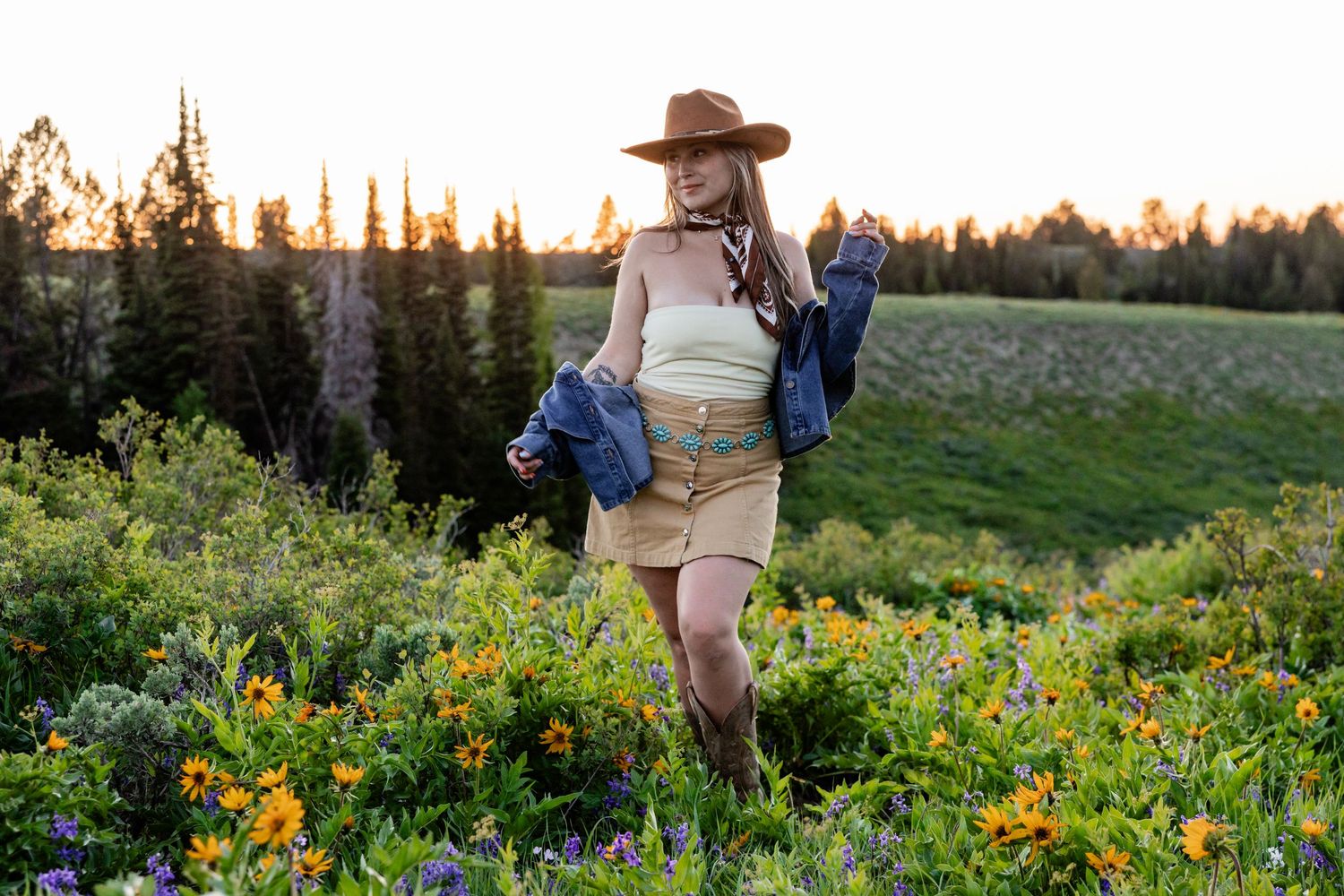 Woman in cowboy hat, tan skirt, and boots poses in wildflower meadow at golden hour with forest backdrop.