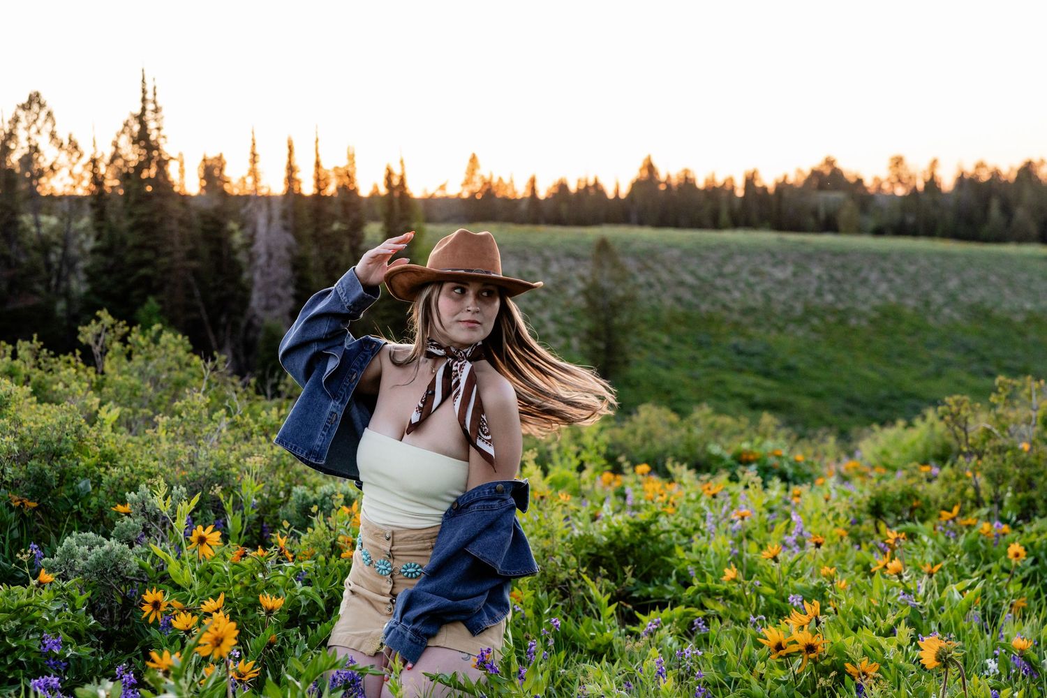 Woman in cowboy hat and denim jacket poses among wildflowers in a mountain meadow at golden hour sunset.