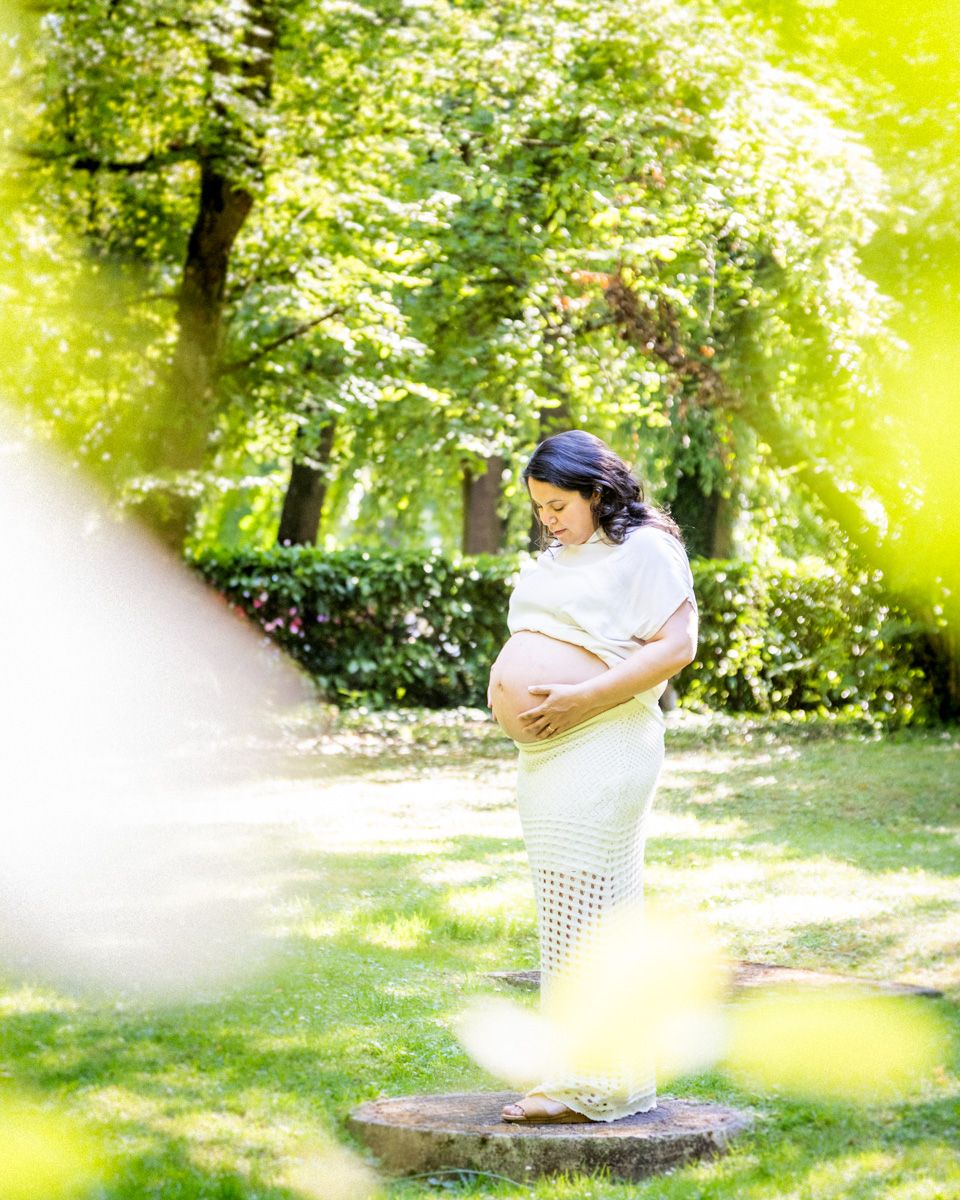 Pregnant woman in white dress cradling baby bump in sunlit park with lush green trees.