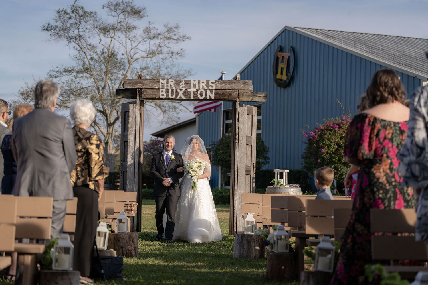 Bride and her father entering the ceremony under a wooden arch with the Blue Barn behind them at a Rockin' H Ranch wedding in Palm City, FL