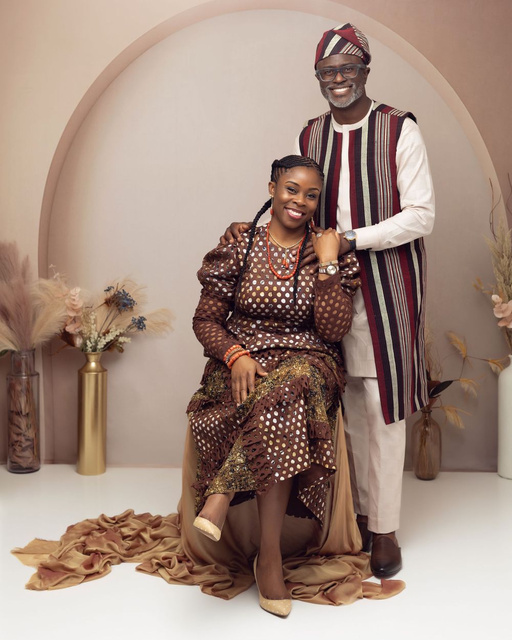 Couple in matching brown African print outfits posing elegantly in a studio with arched backdrop.