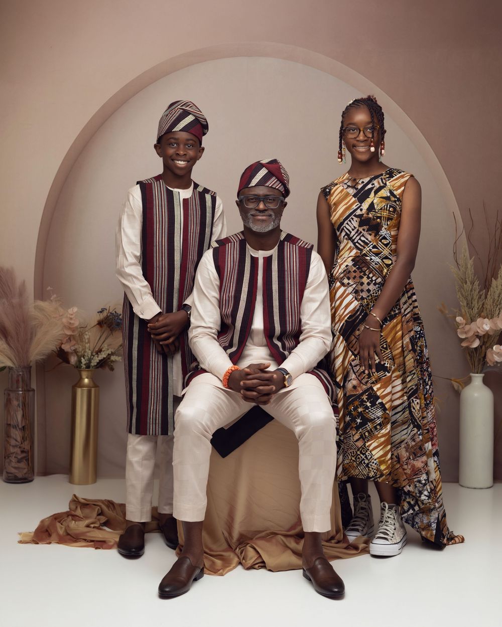 Three people in colorful African traditional attire pose for a portrait in a studio with arch backdrop.