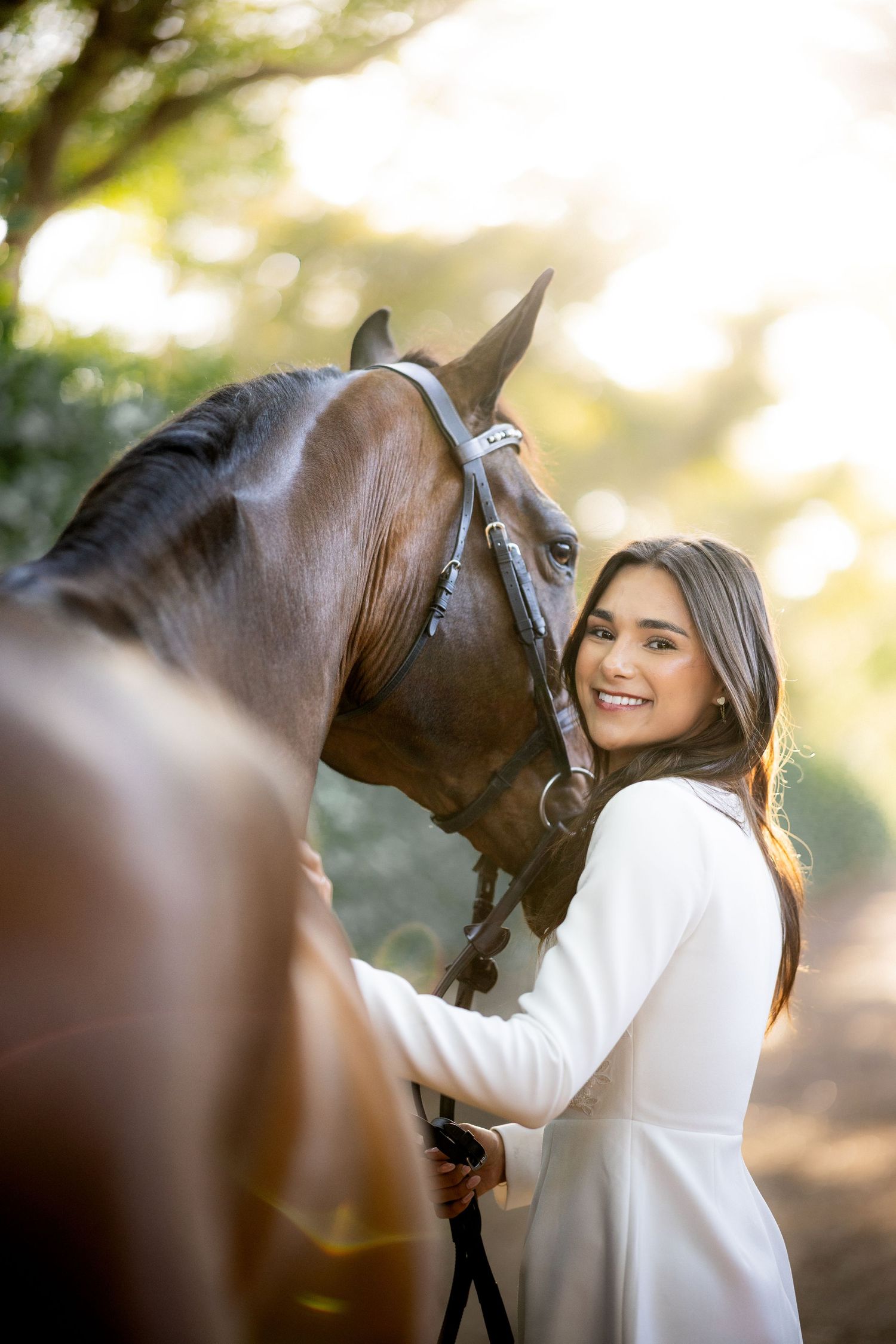 Young woman in white dress smiling beside a brown horse on a sunlit tree-lined path.