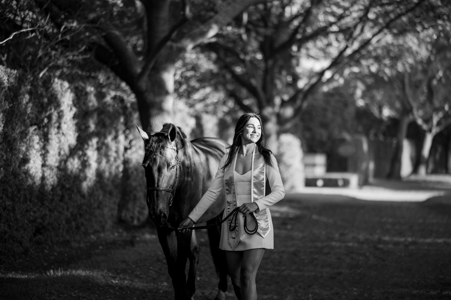 Black and white photo of a smiling woman standing beside a horse on a tree-lined path.